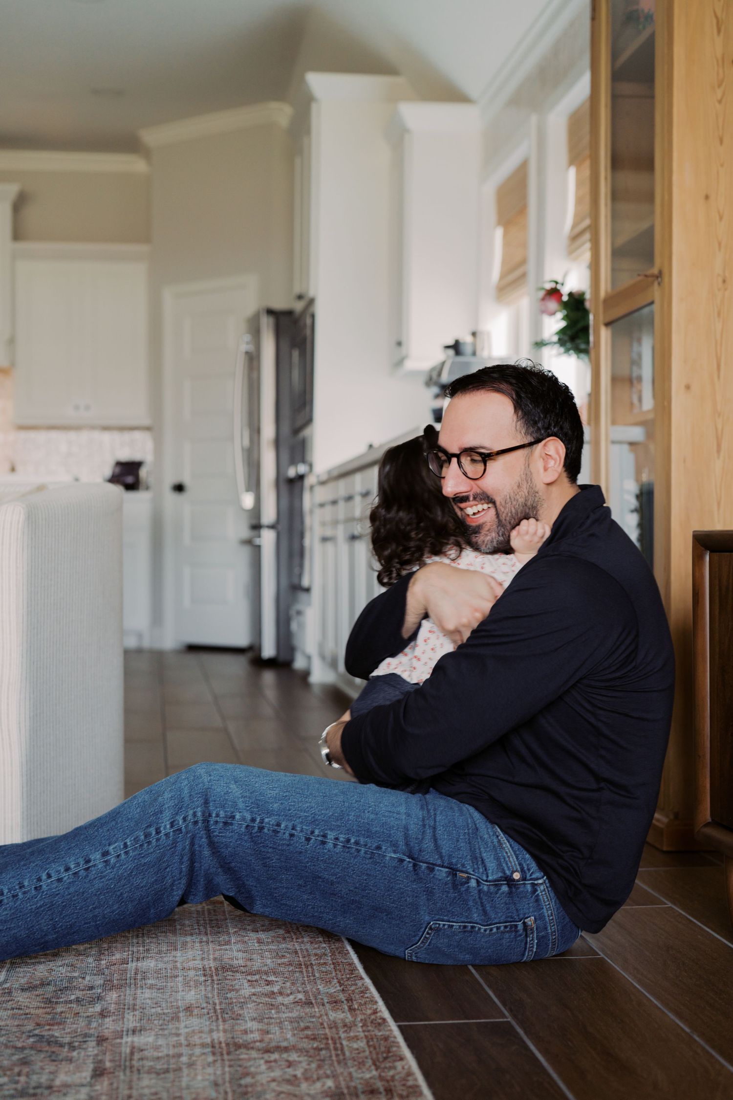 A man with curly hair sits on the floor of a home, embracing a woman who is smiling and leaning against him.