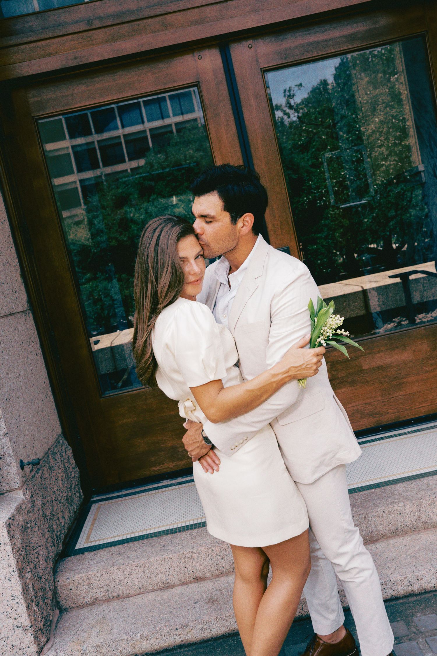 A couple in matching white outfits embrace and kiss on stone steps outside a building with large wooden doors.