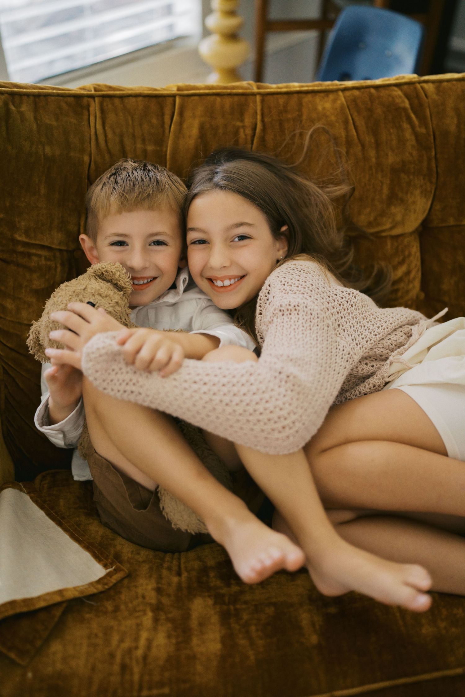 Two happy children cuddle together on a brown velvet armchair, smiling with a teddy bear.
