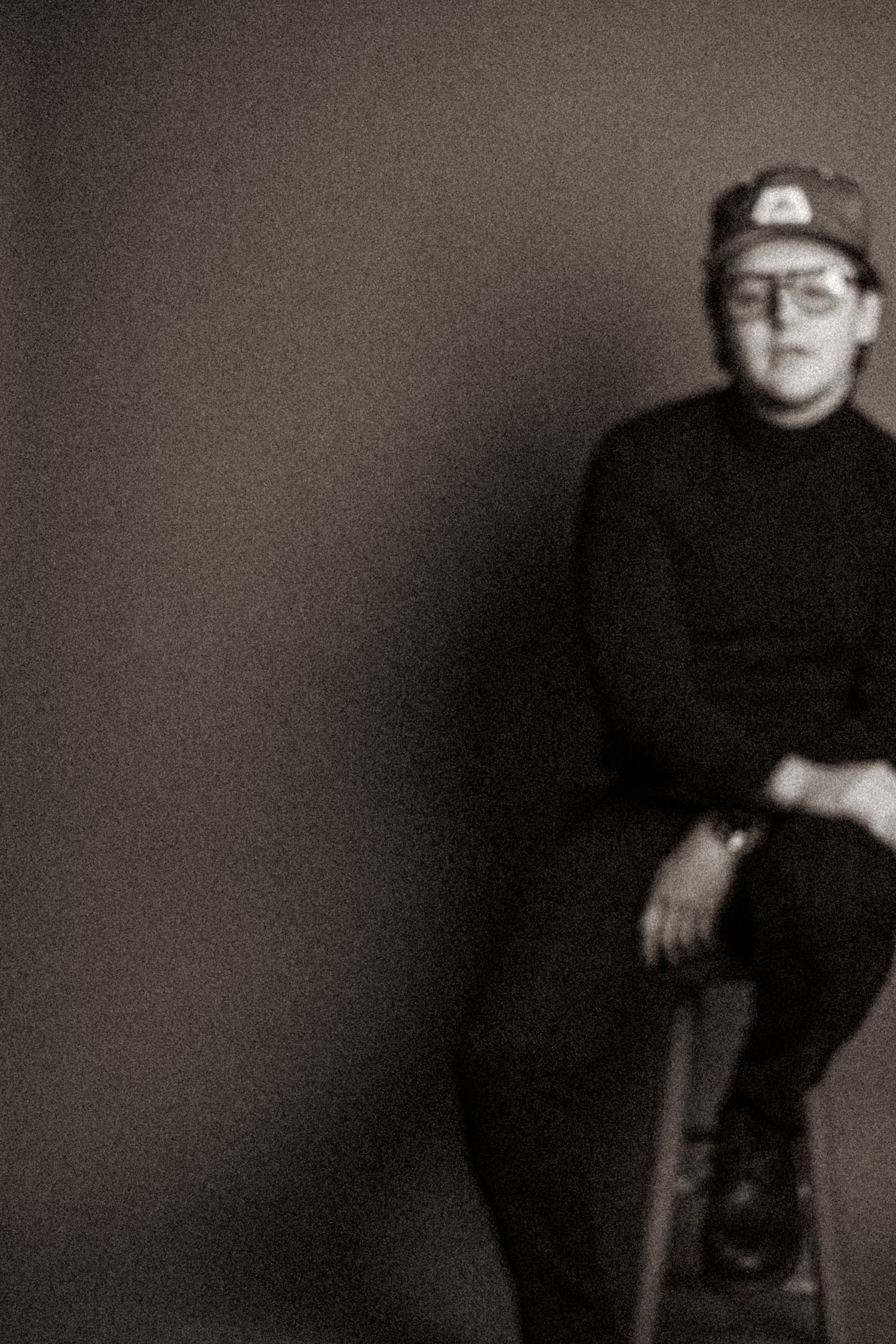 Black and white portrait of person in cap and glasses sitting on stool in studio.