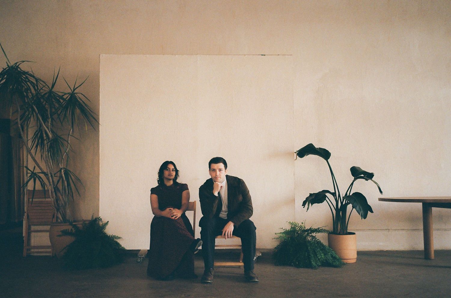 Couple sitting casually in minimalist studio space surrounded by indoor plants on wooden floor.