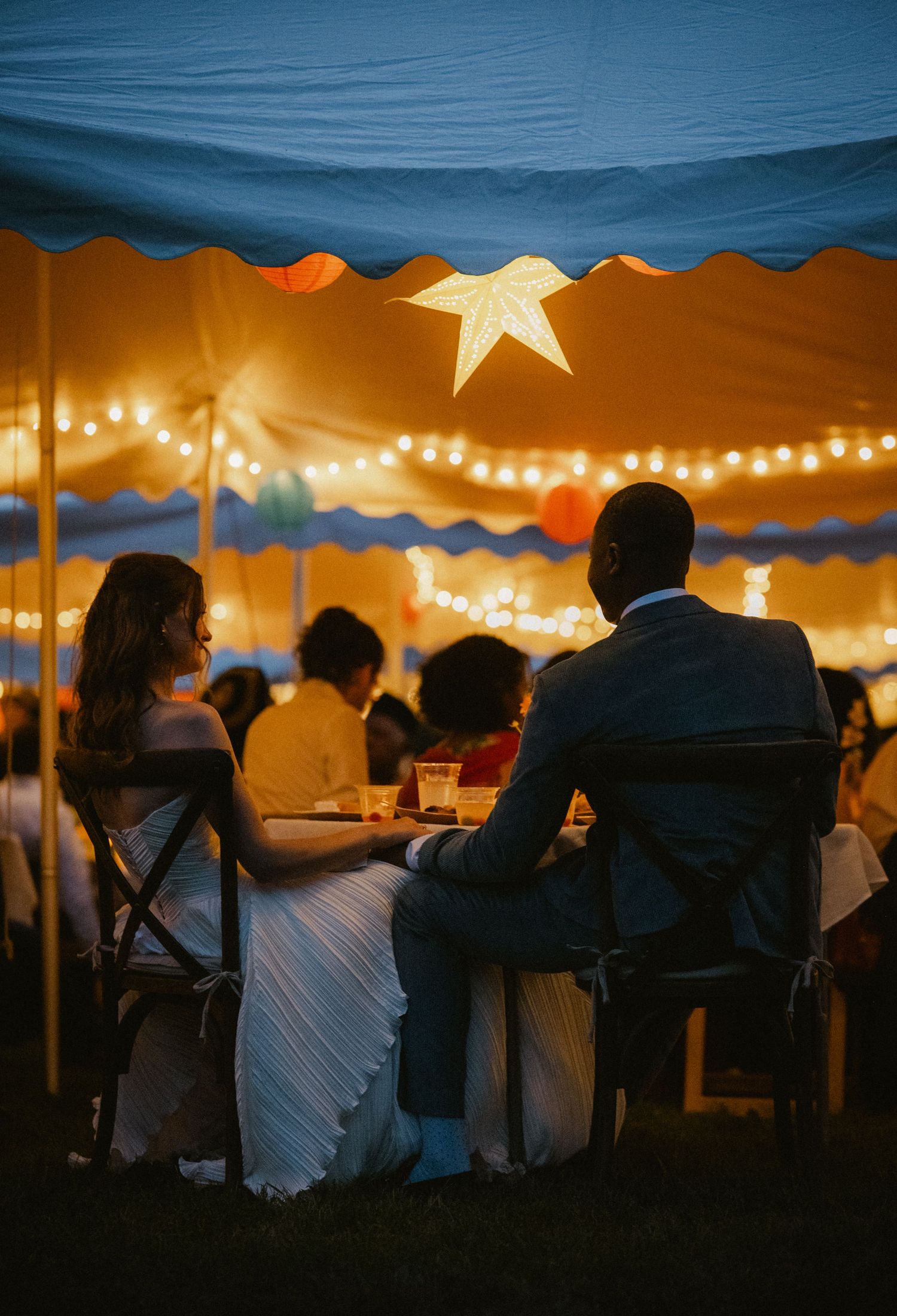 Couple seated at candlelit wedding reception under a glowing tent with string lights and a star decoration.