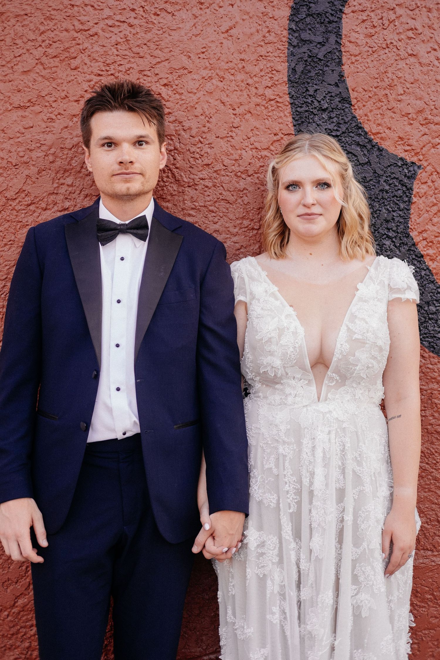 Bride and groom holding hands in front of a red wall, wearing a white lace gown and navy tuxedo.