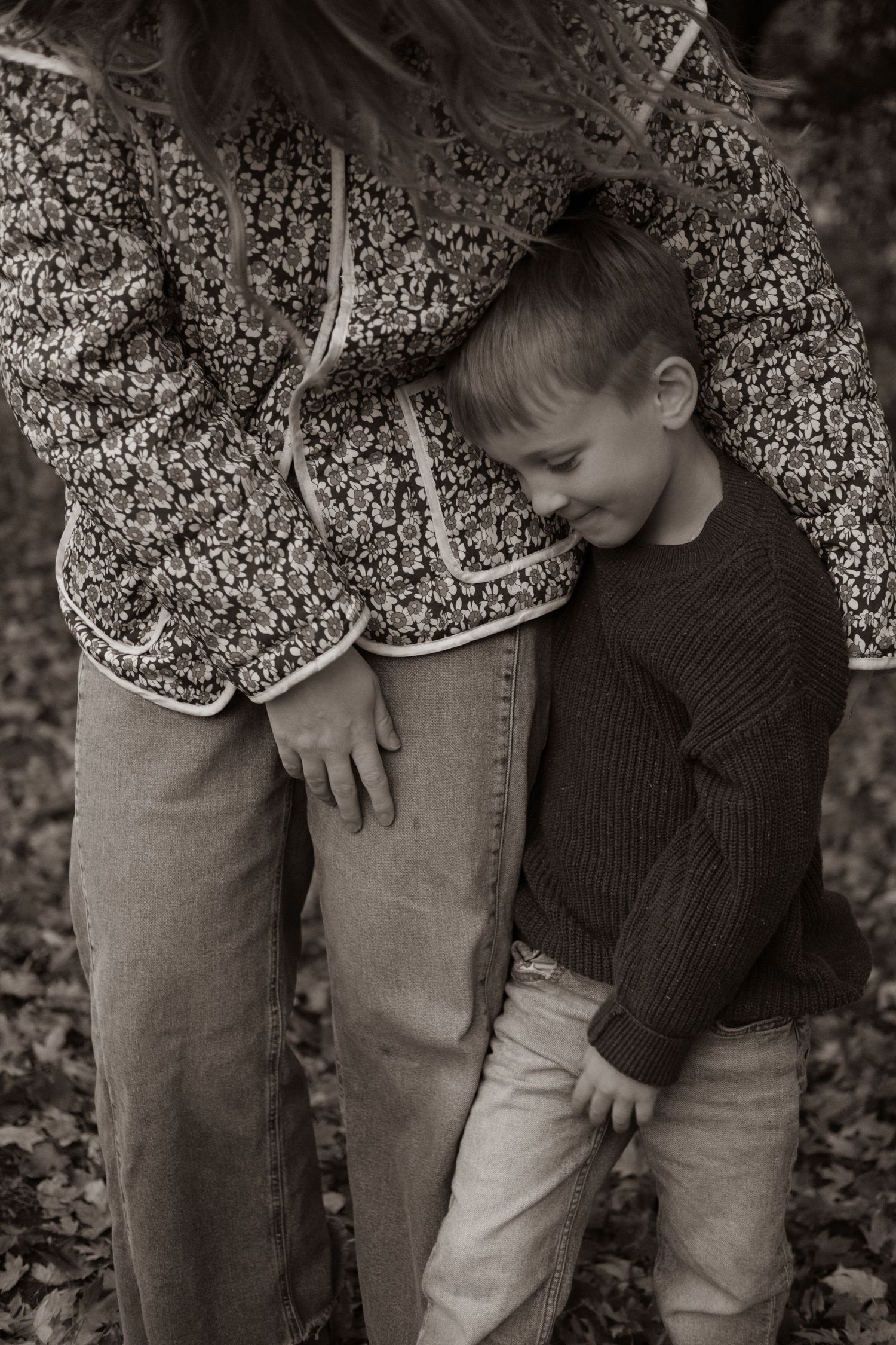 Black and white photo of a young boy leaning against his mother's side outdoors among autumn leaves.