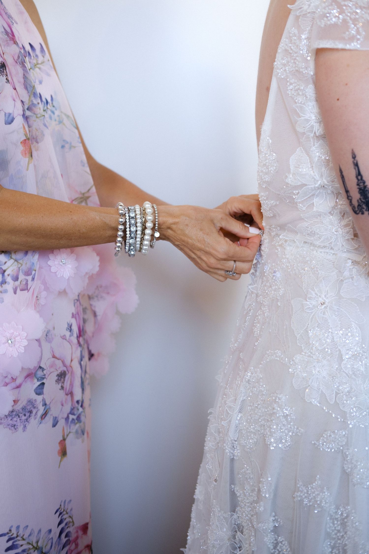 A woman in floral dress fastens buttons on a bride's embellished white wedding gown, wearing stacked crystal bracelets.