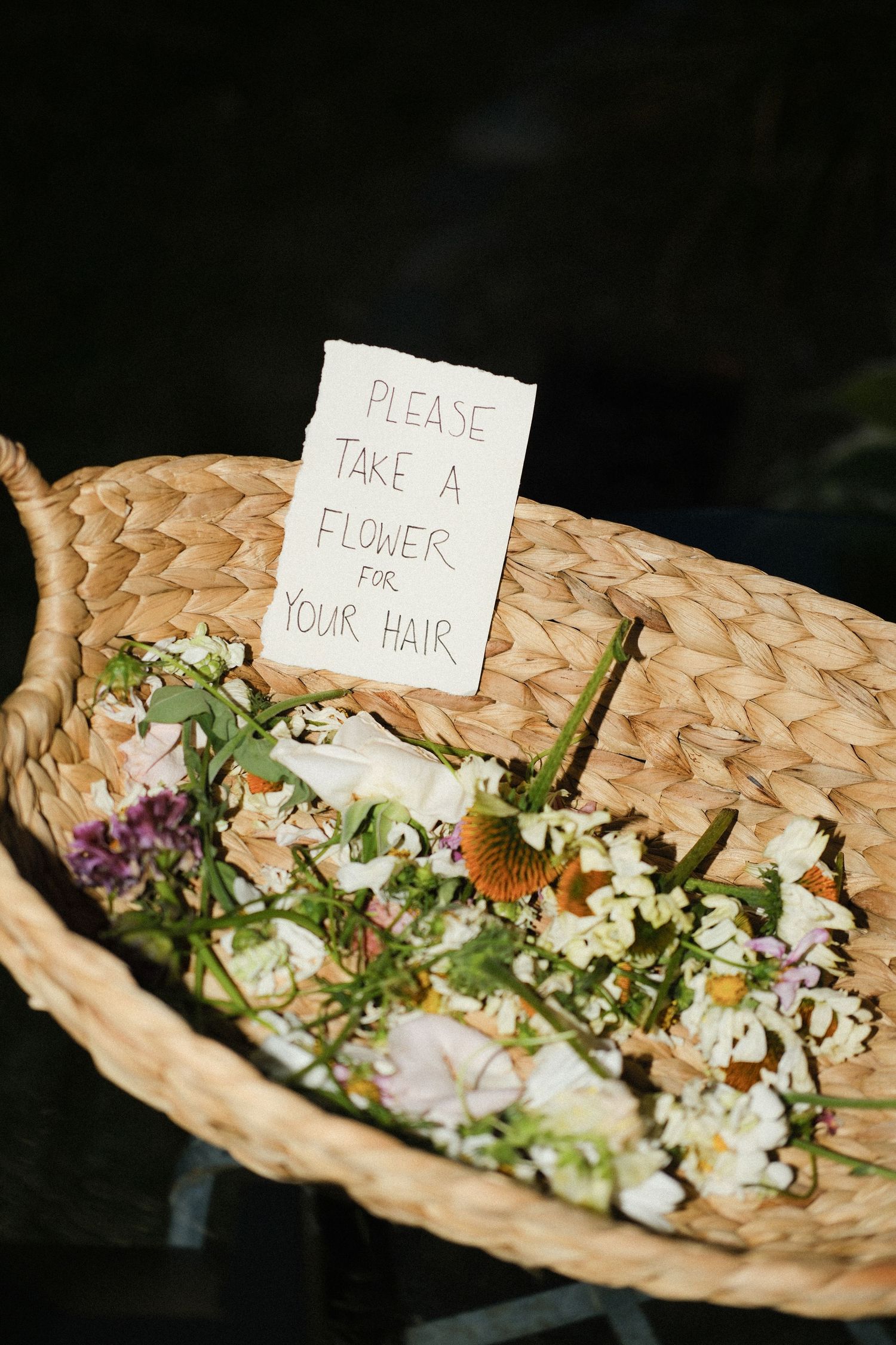 Wicker basket filled with wildflowers and a handwritten sign reading Please Take a Flower for Your Hair at a wedding.