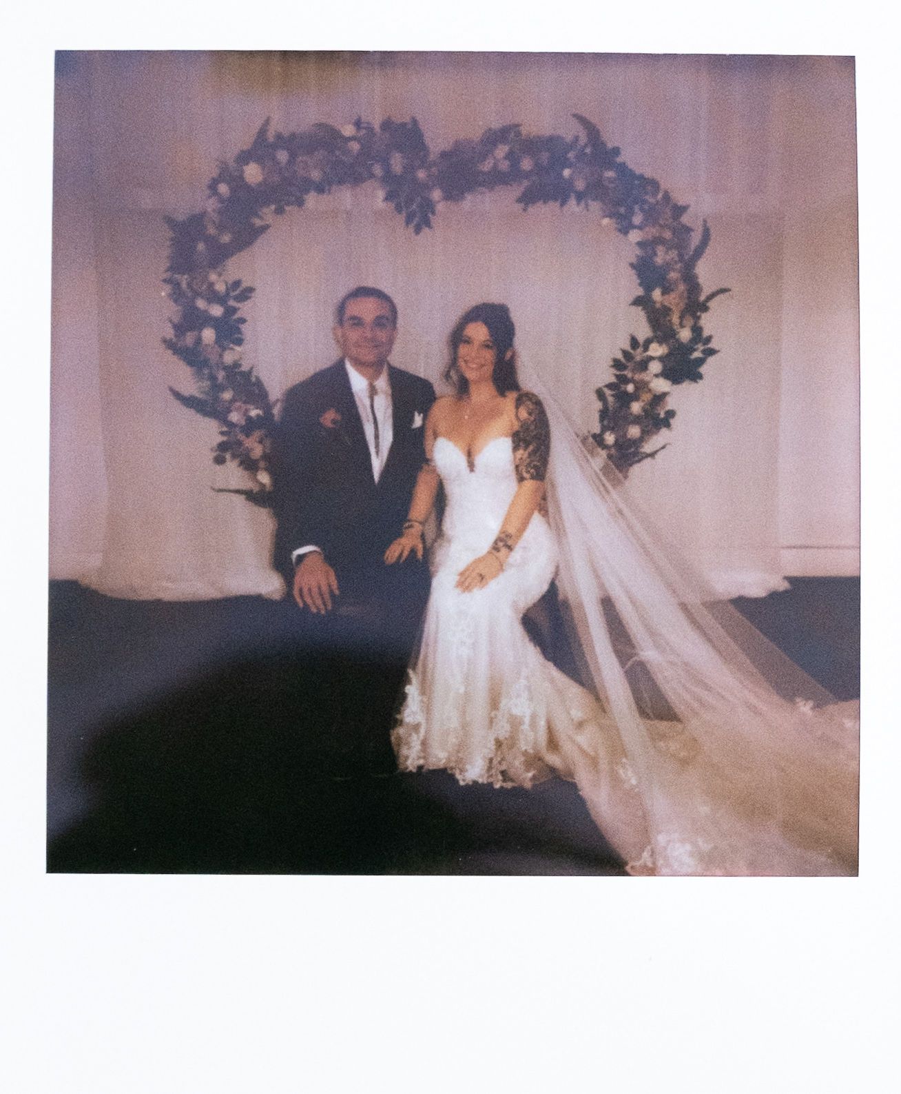 A bride and groom pose together under a floral heart arch in a classic wedding portrait captured on Polaroid film.