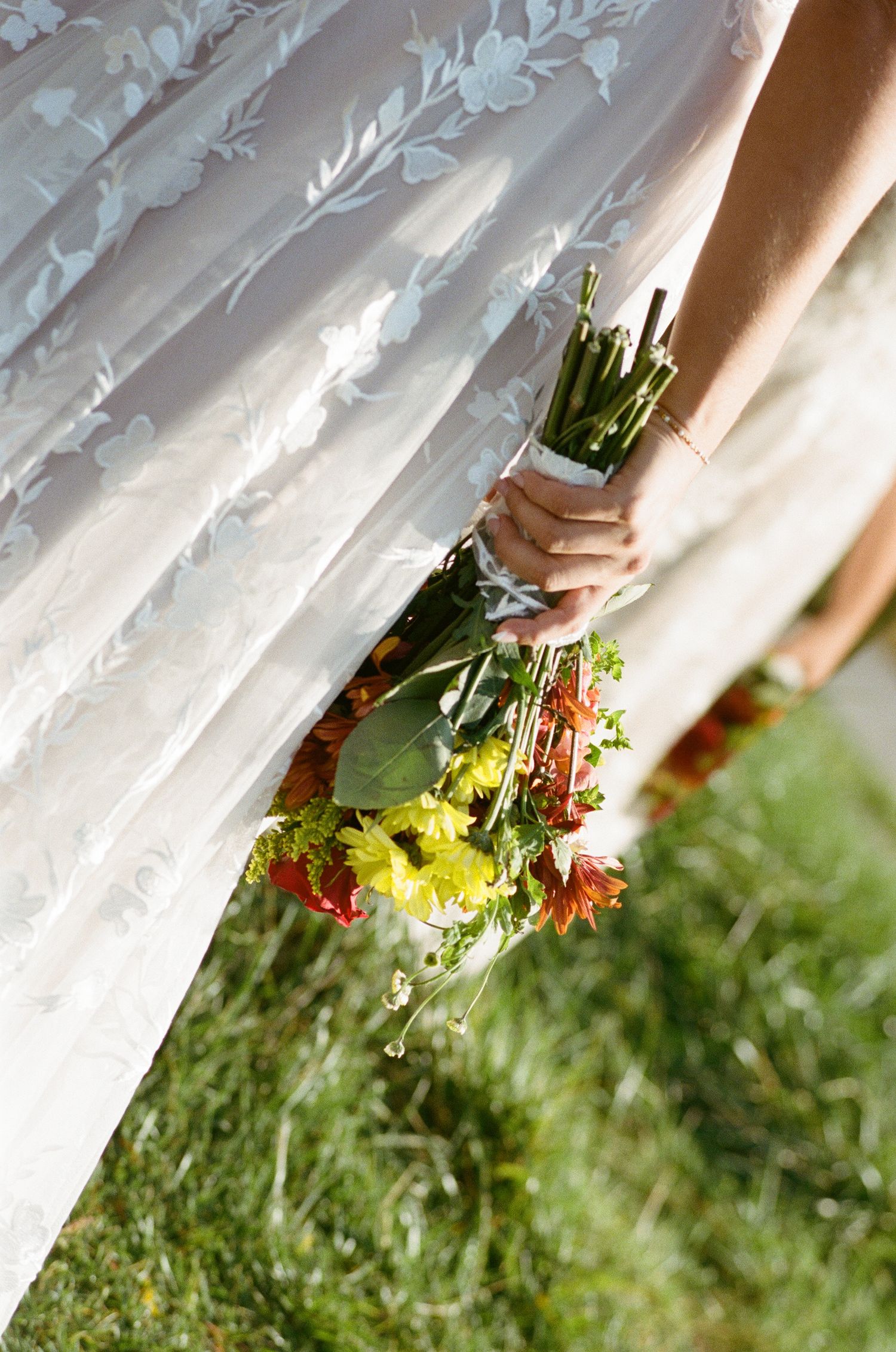 Bride in floral lace gown holding colorful yellow and orange wildflower bouquet outdoors.