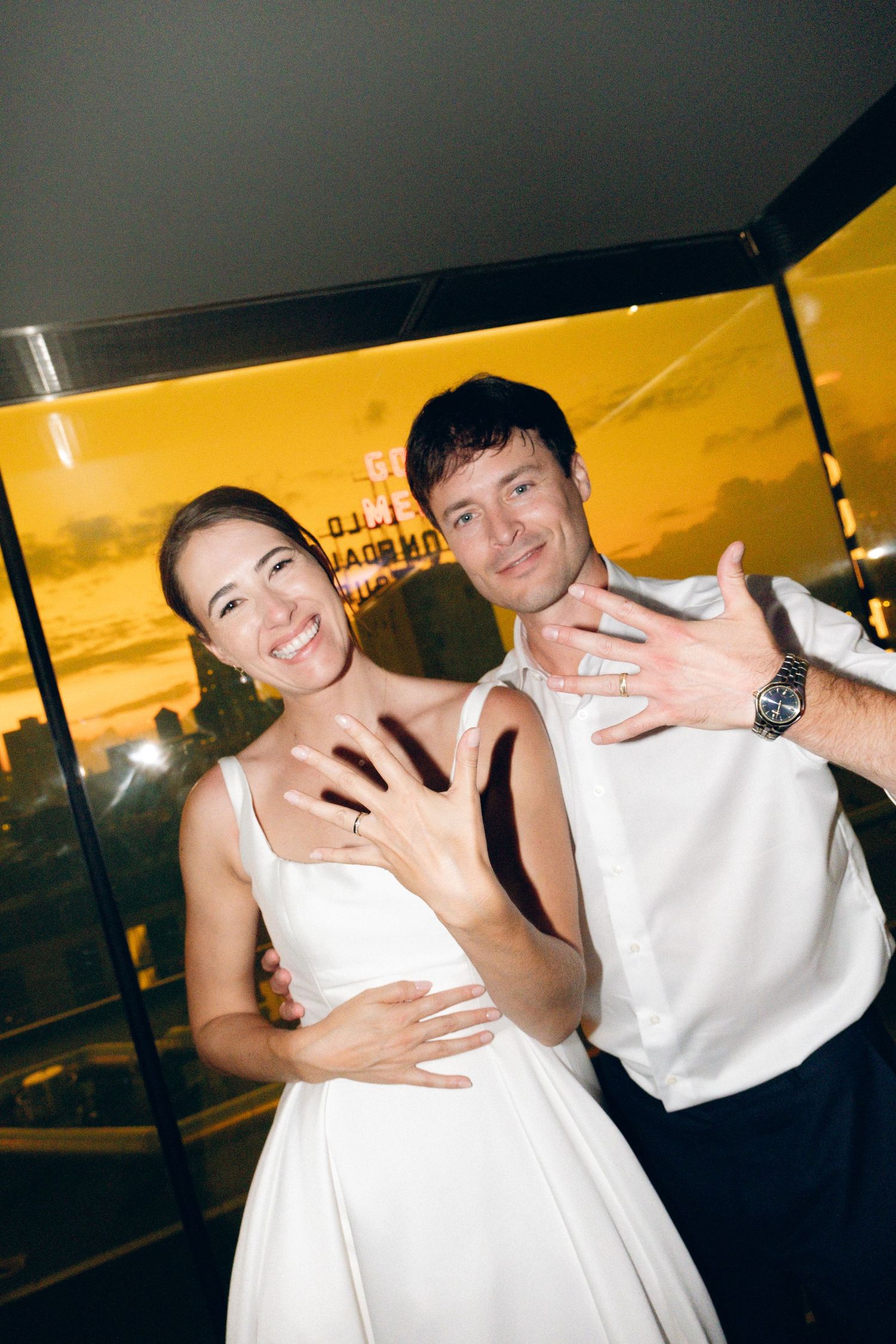 Happy newlyweds showing off wedding rings, smiling couple in white attire at golden sunset venue.