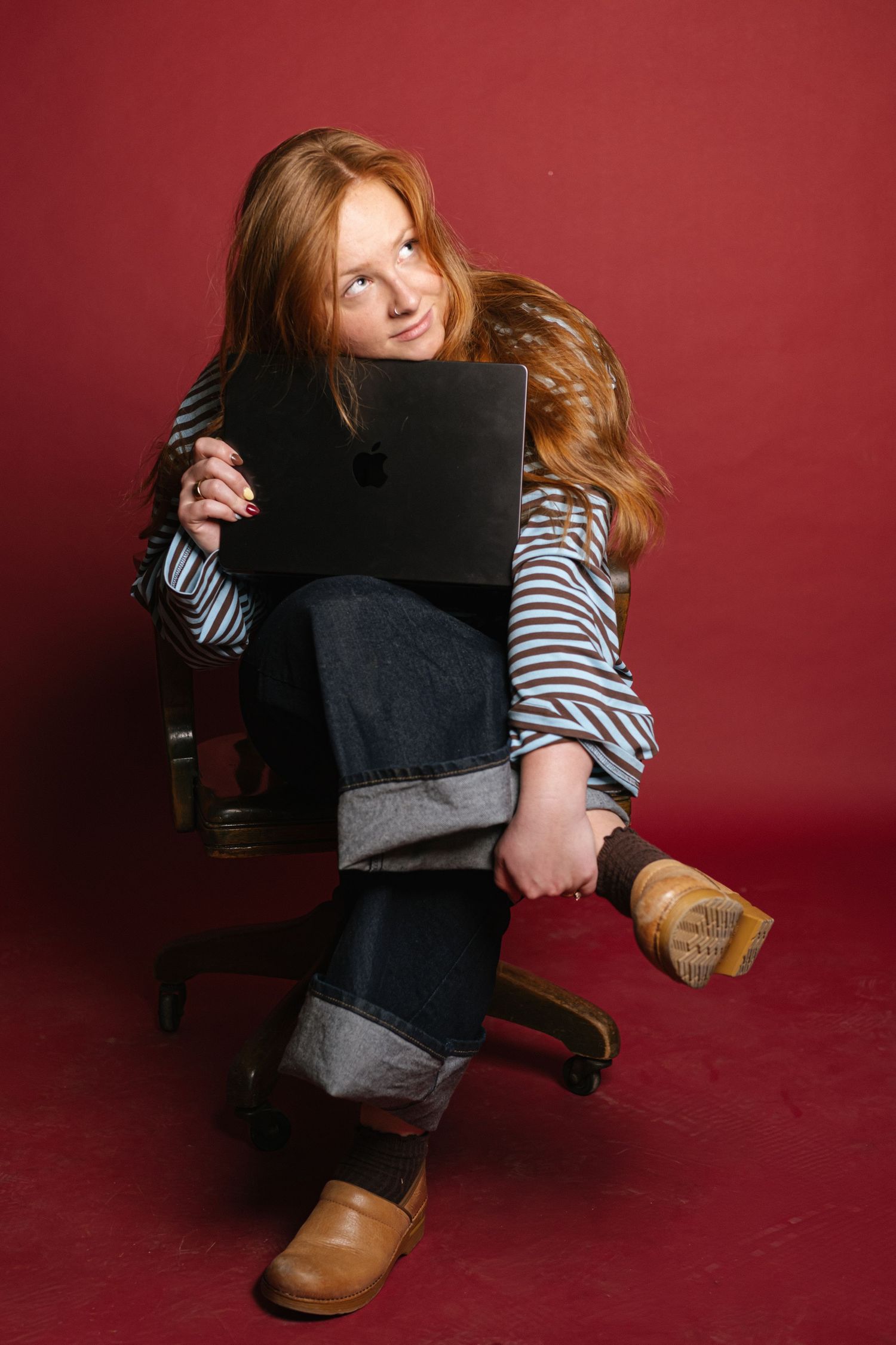Red-haired girl sitting in office chair hugging Apple MacBook laptop against red background.