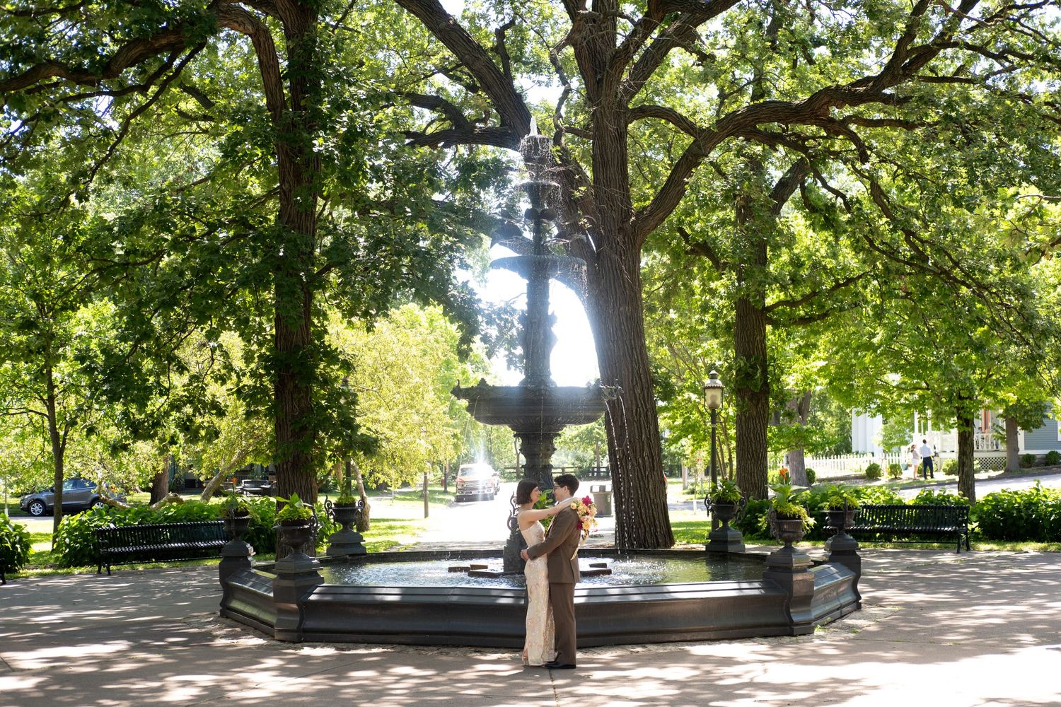 Couple embracing by a decorative fountain surrounded by lush green trees in a sunny park.