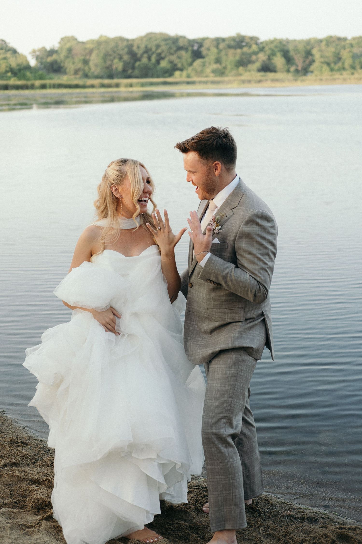 Joyful bride and groom laughing together by a lakeside, she in a white gown, he in a gray suit.