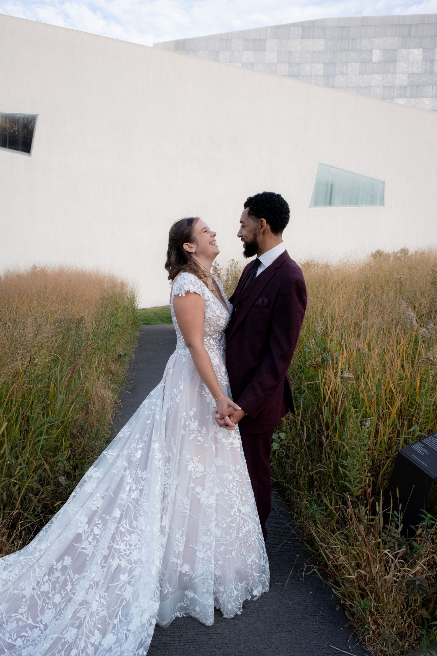 Newlywed couple in wedding attire holding hands and smiling at each other outdoors.