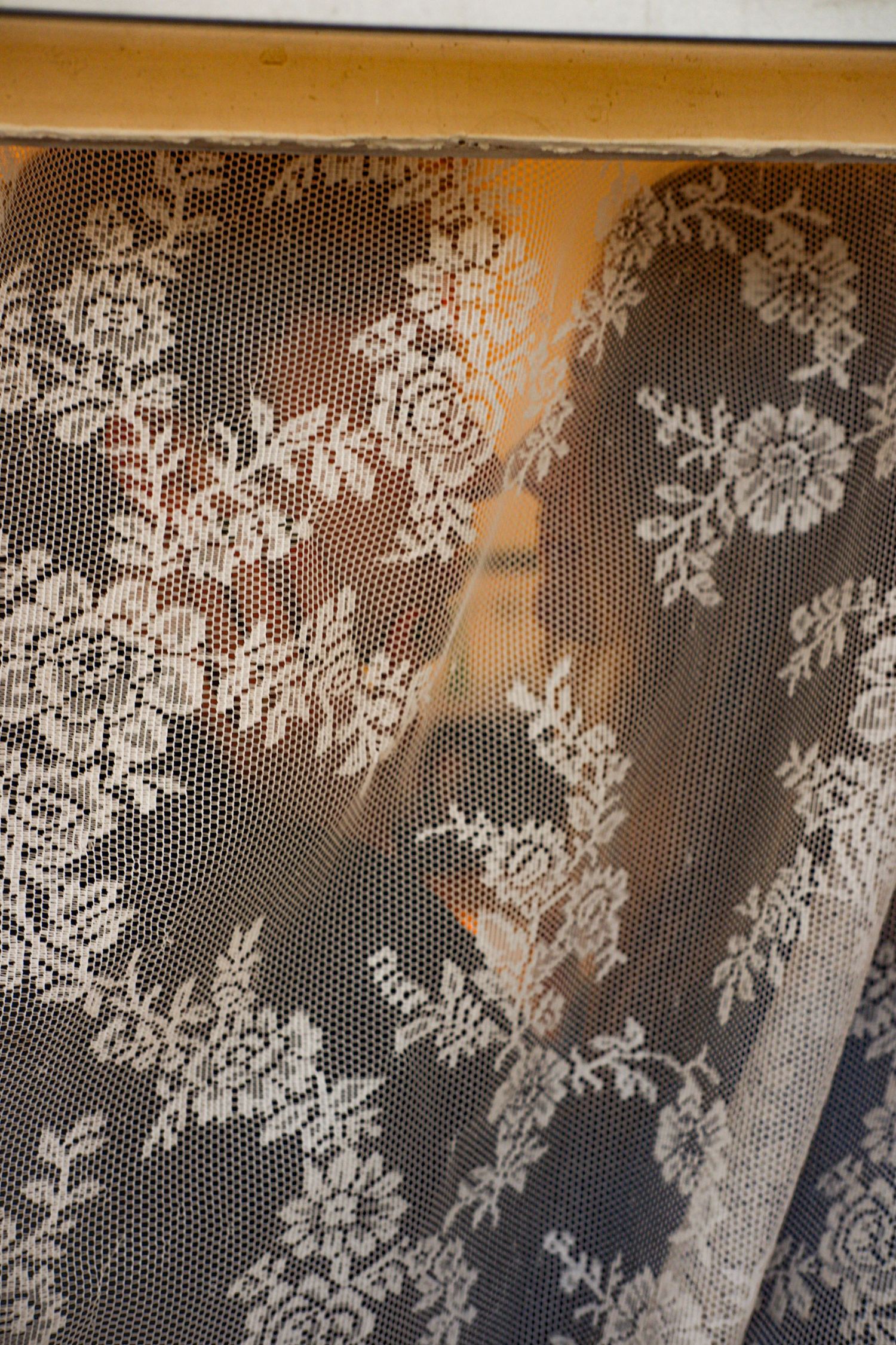 Delicate white lace curtain with floral pattern softly diffusing warm light through window.