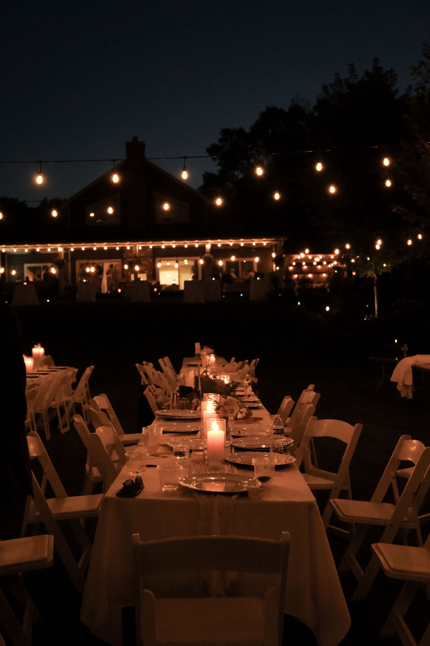 Elegant outdoor evening wedding reception with string lights, candlelit tables, white chairs, and a glowing venue in the background.