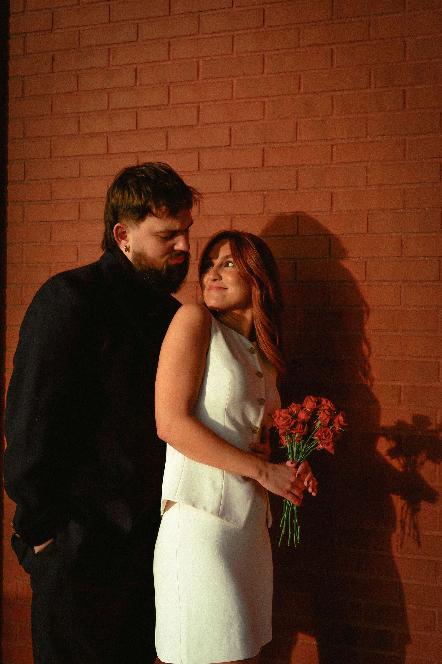 Couple embracing against brick wall in warm light, bride holding red flowers.