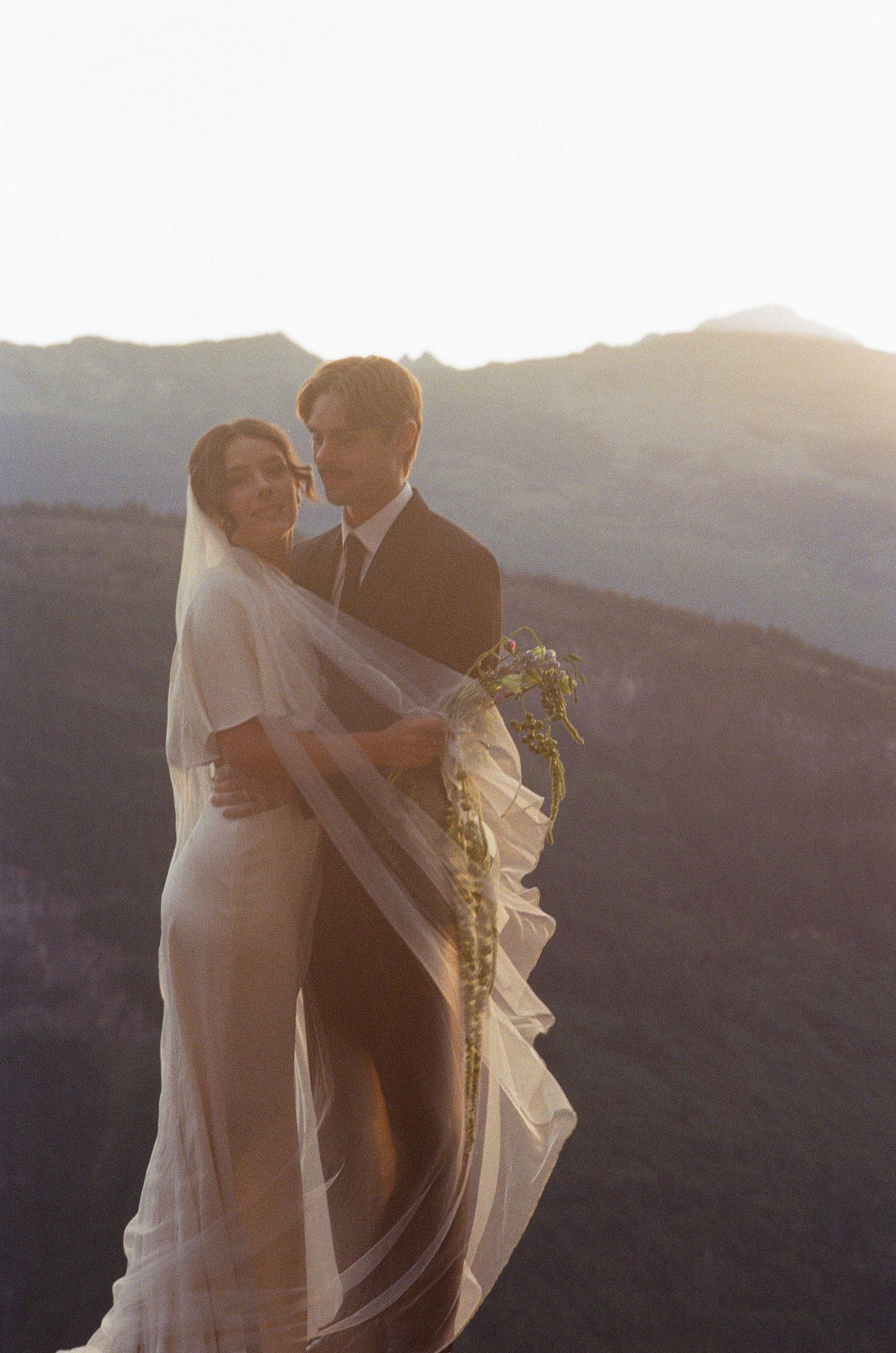 Bride and groom embracing at sunset on mountain overlook with flowing wedding veil.