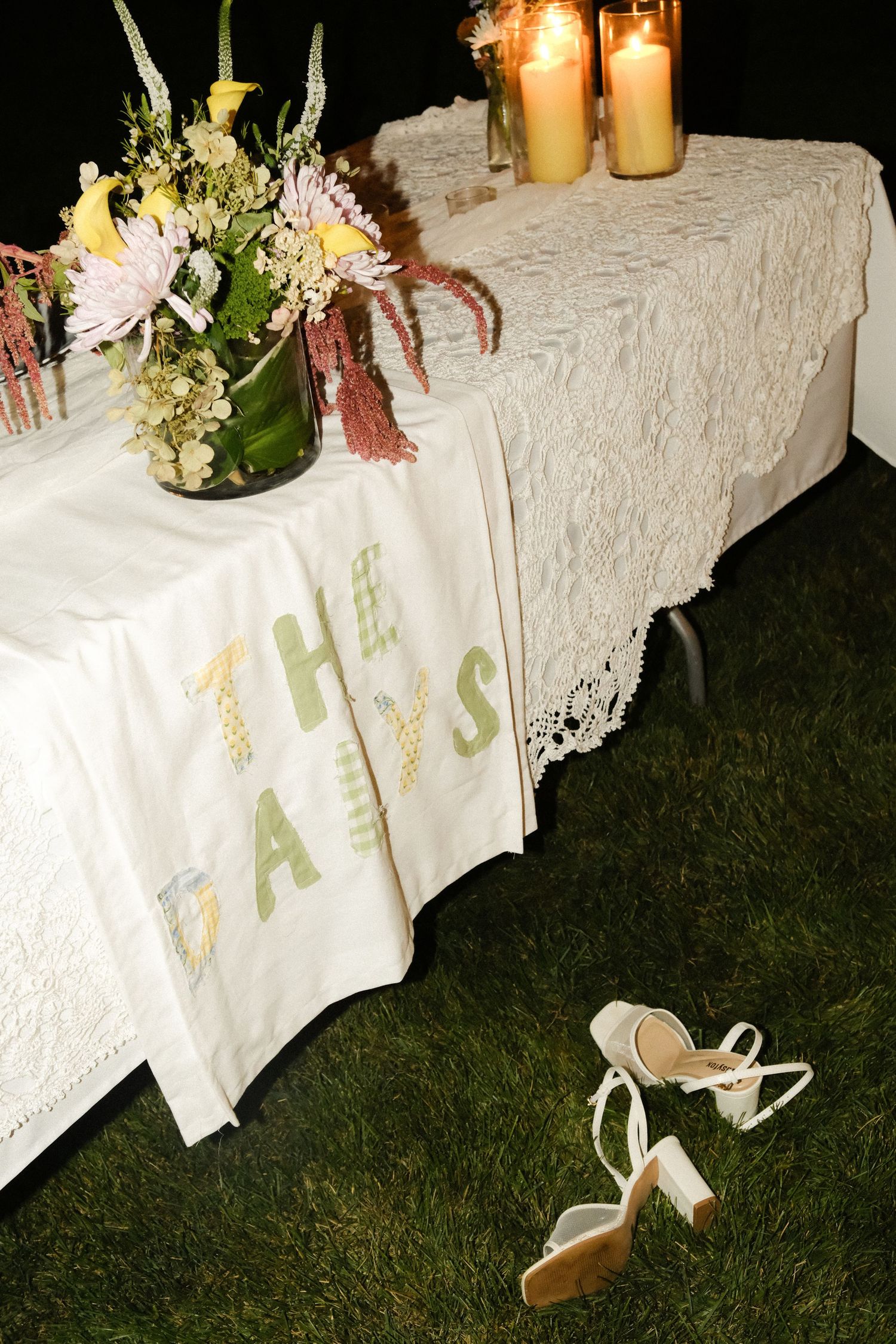 Elegant outdoor wedding sweetheart table with lace cloth, floral arrangement, candles, and white heels on grass at night.