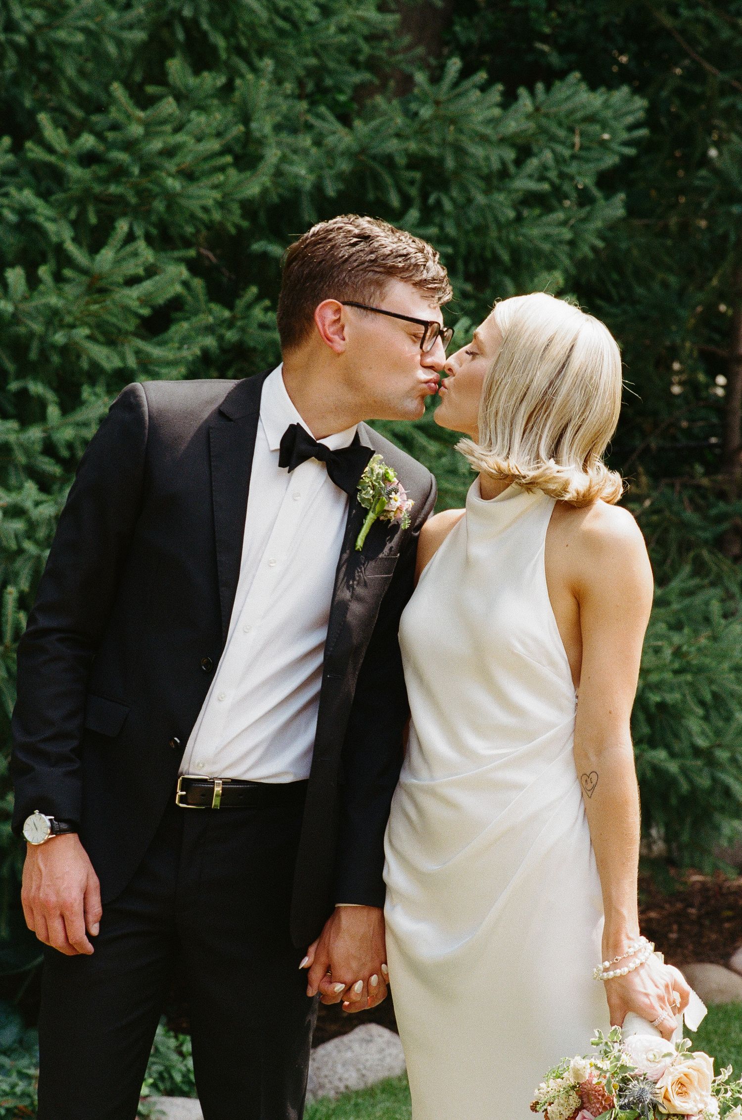 Groom in black tuxedo kissing blonde bride in white dress outdoors near evergreen trees.