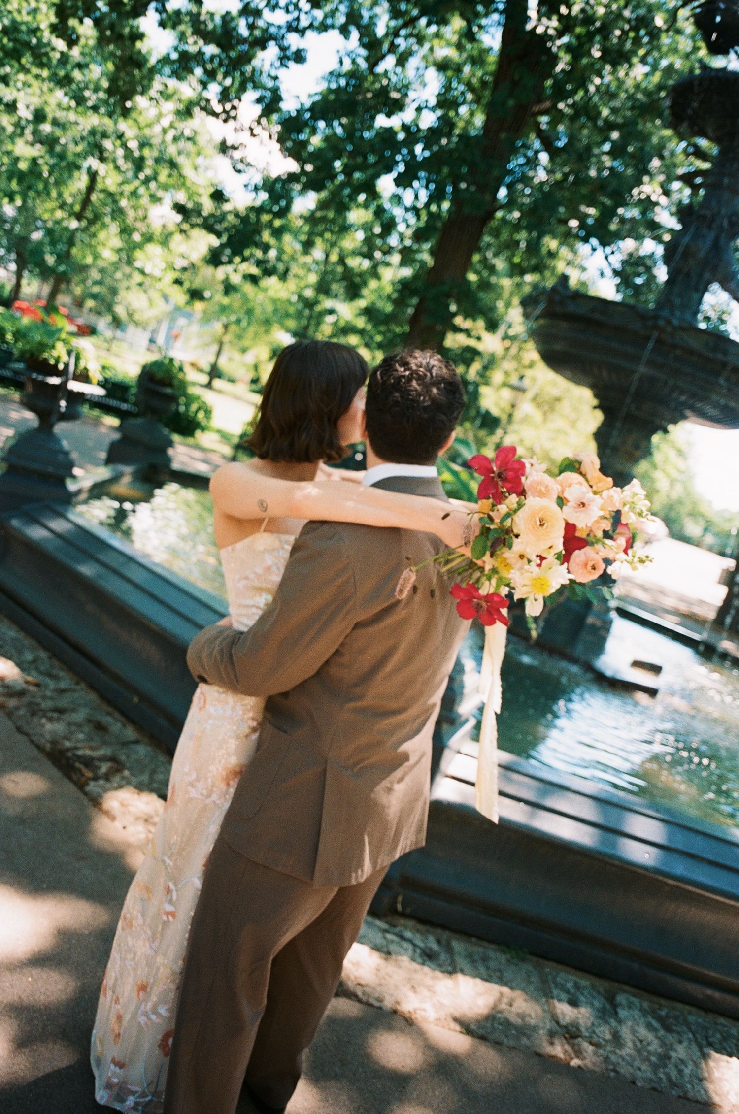 Newlyweds embracing near a park fountain, bride holding a colorful bouquet of pink and orange blooms.