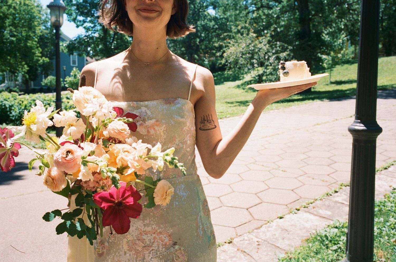 Bride in floral slip dress holding a large colorful bouquet and a slice of cake in a sunny park.