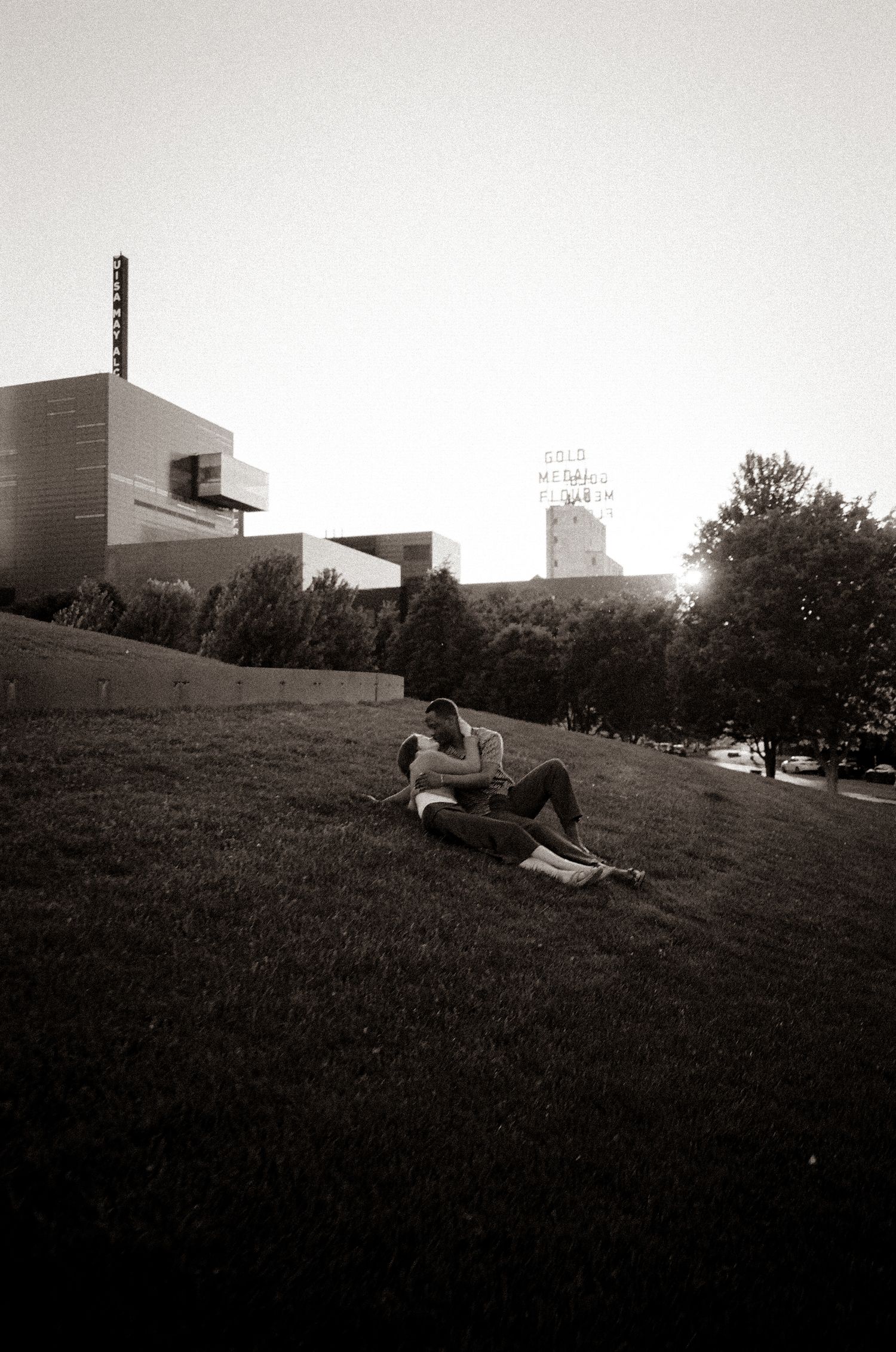 Couple sitting together on a grassy hill near a modern building, captured in black and white film photography.