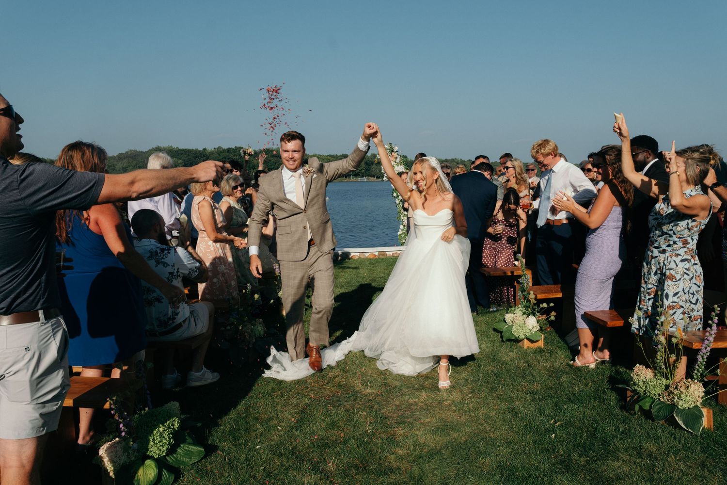 Bride and groom walk triumphantly down the aisle outdoors, guests cheering, with a scenic waterfront backdrop.