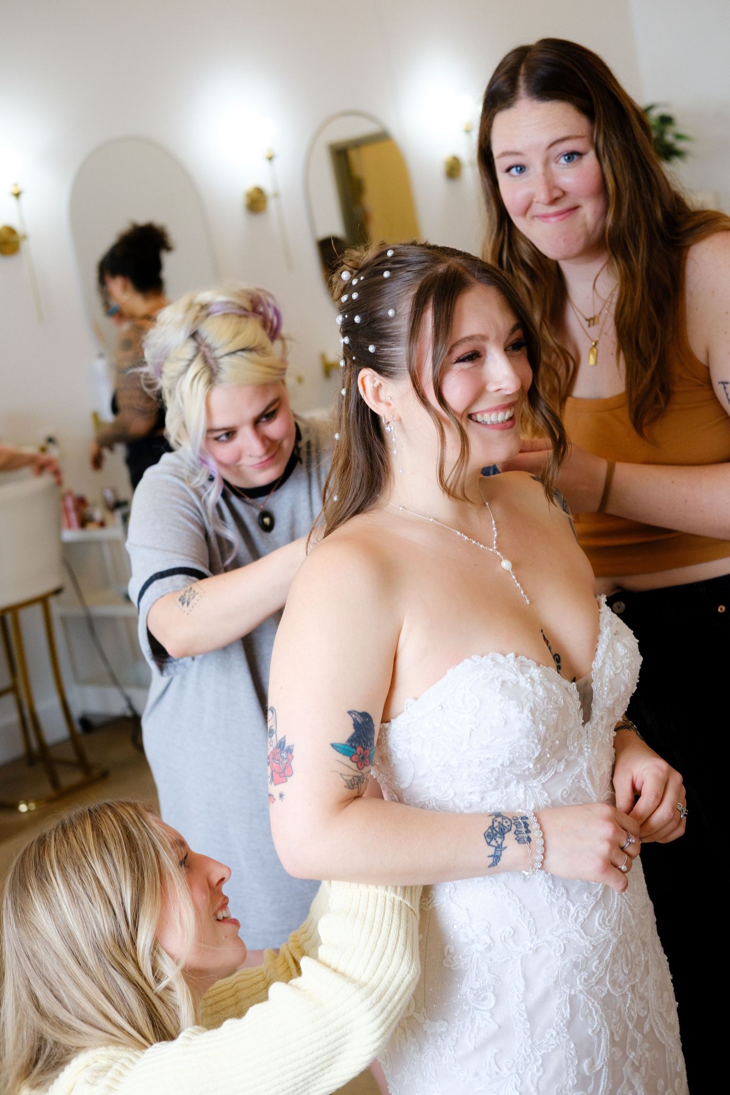 Bridesmaids helping smiling bride into white wedding dress in bright salon setting.