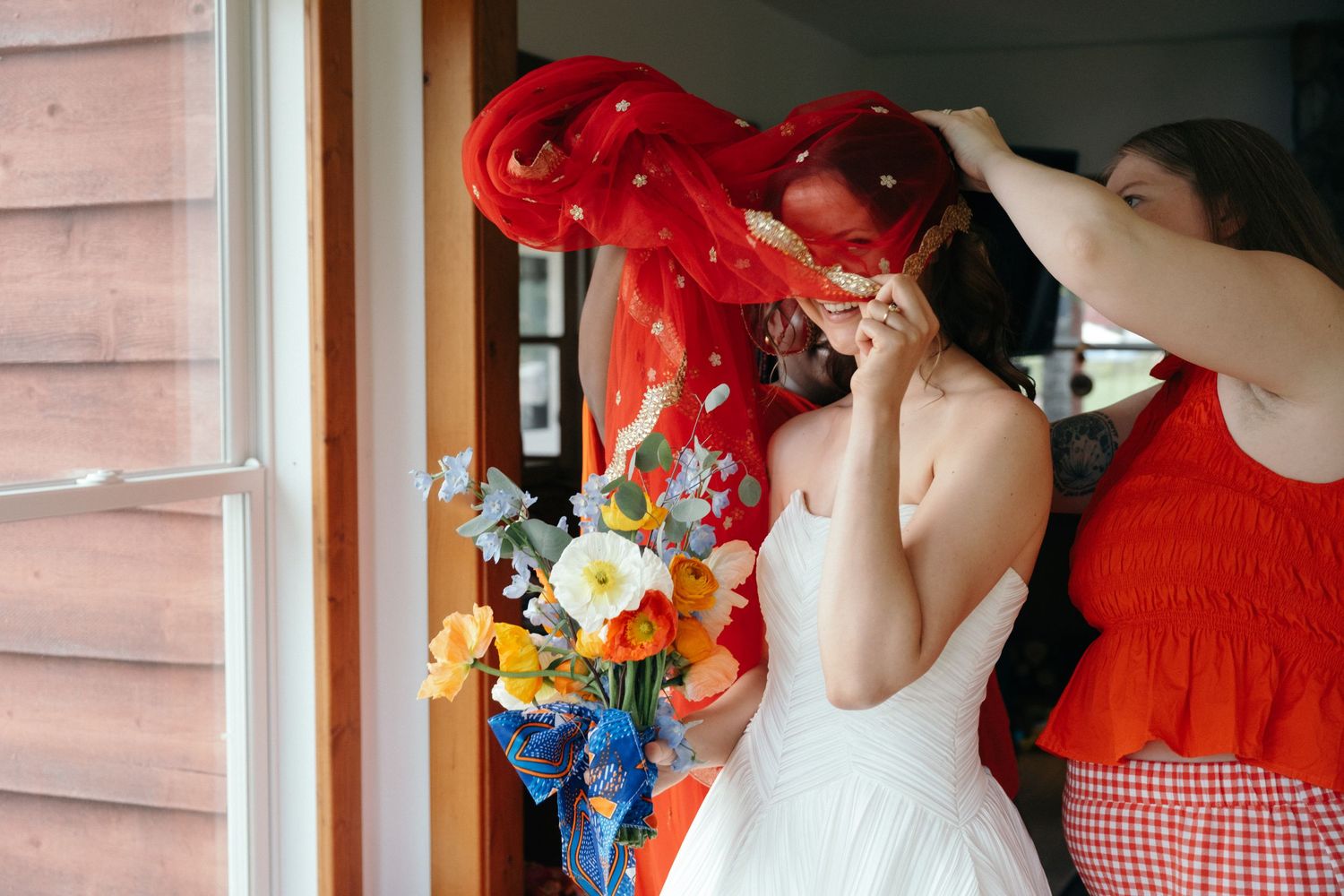 Bride in white dress with red veil being adjusted by bridesmaid, colorful bouquet visible in background.
