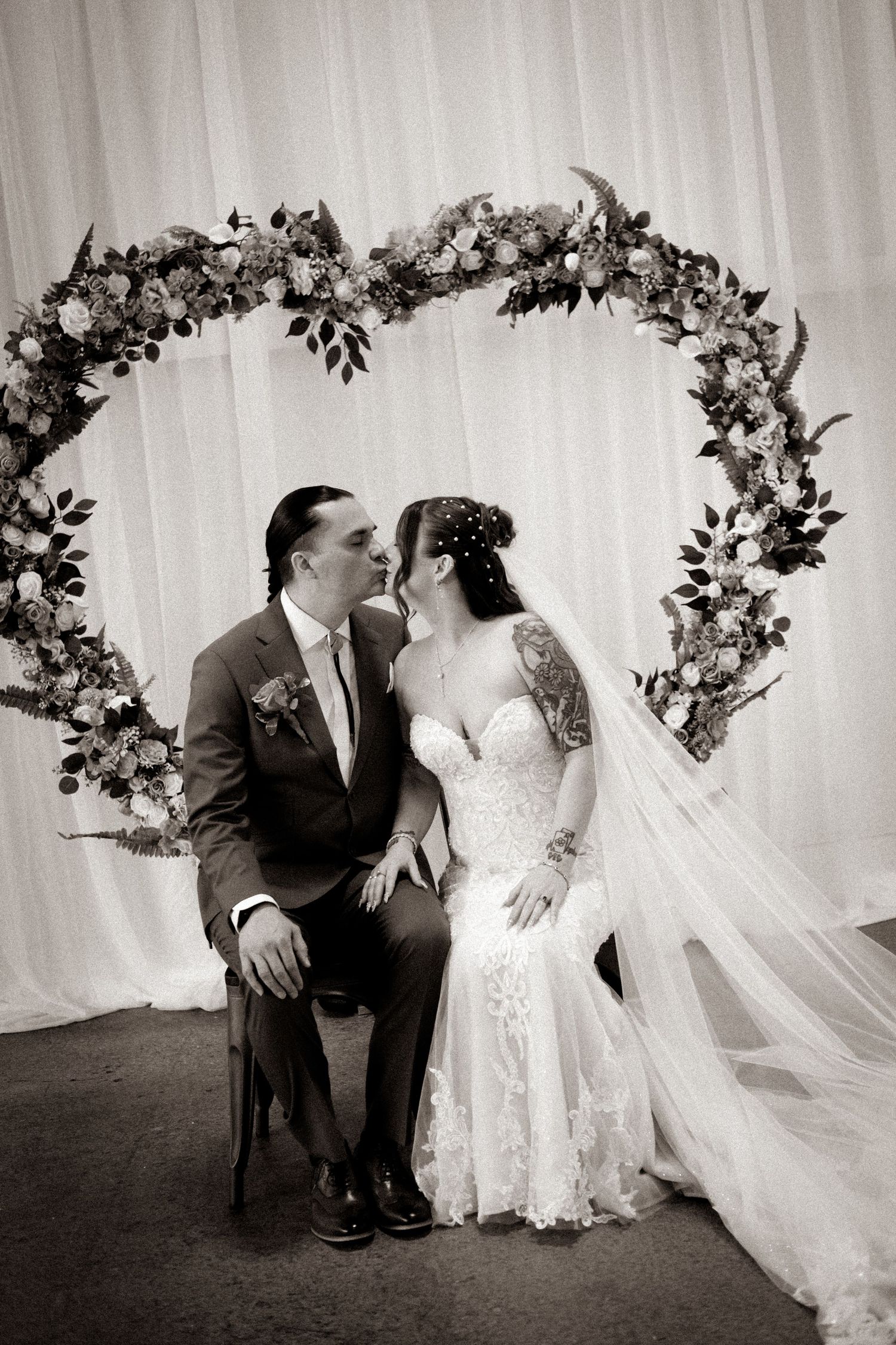 Black and white photo of a bride and groom kissing under a floral heart arch at their wedding ceremony.
