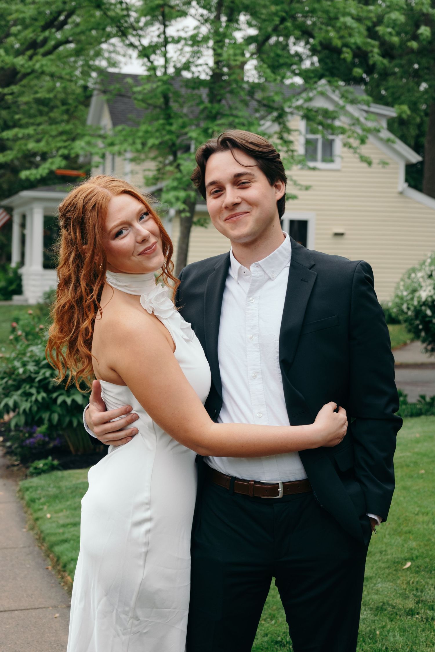 A couple in formal attire embracing outdoors, woman in white dress and man in black suit.