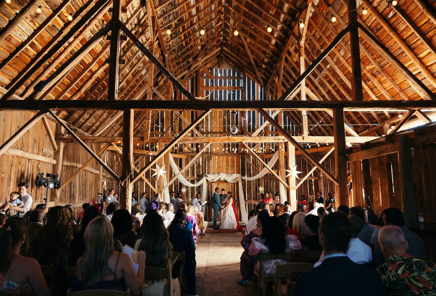 Rustic barn wedding ceremony with wooden beams, string lights, and guests seated as couple stands at the altar.
