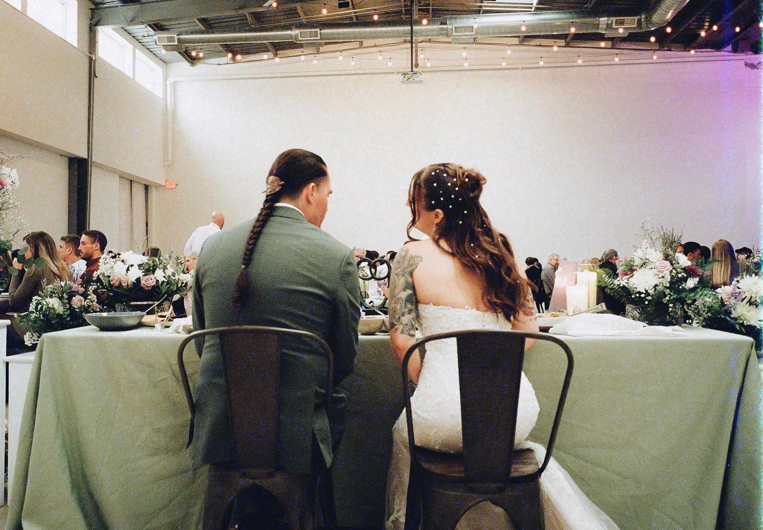 Bride and groom seated at wedding reception table, viewed from behind, surrounded by elegant floral centerpieces.
