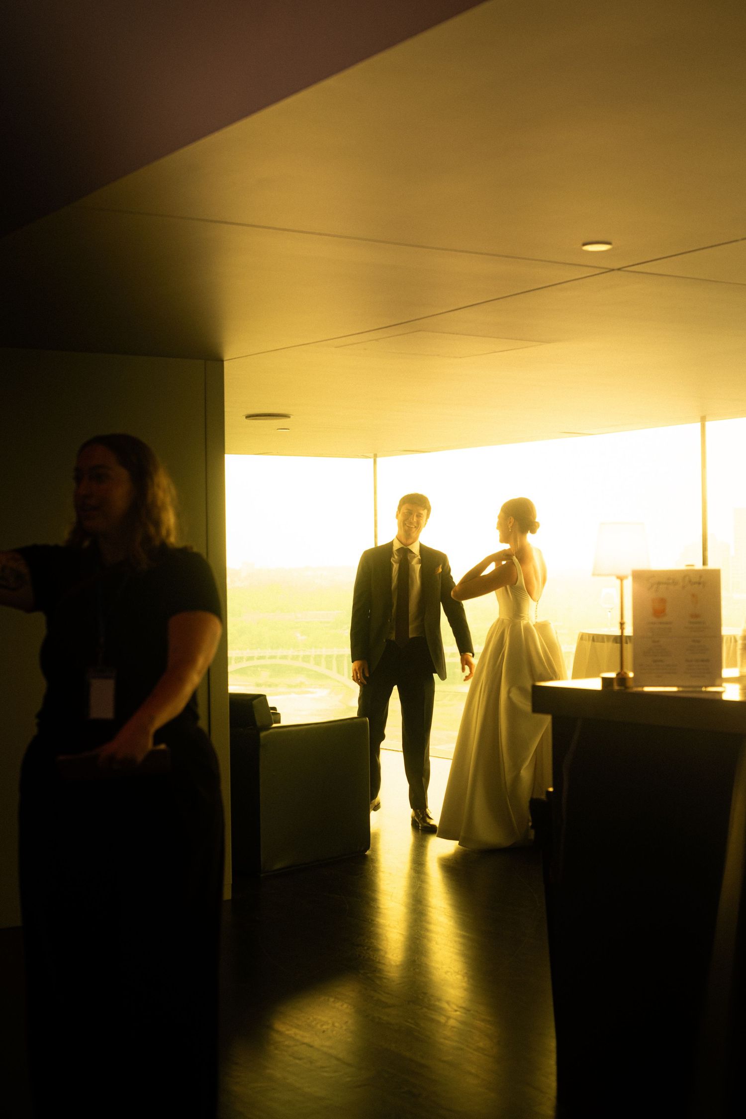 Bride and groom silhouetted against golden sunset light in elegant venue with staff member in foreground.