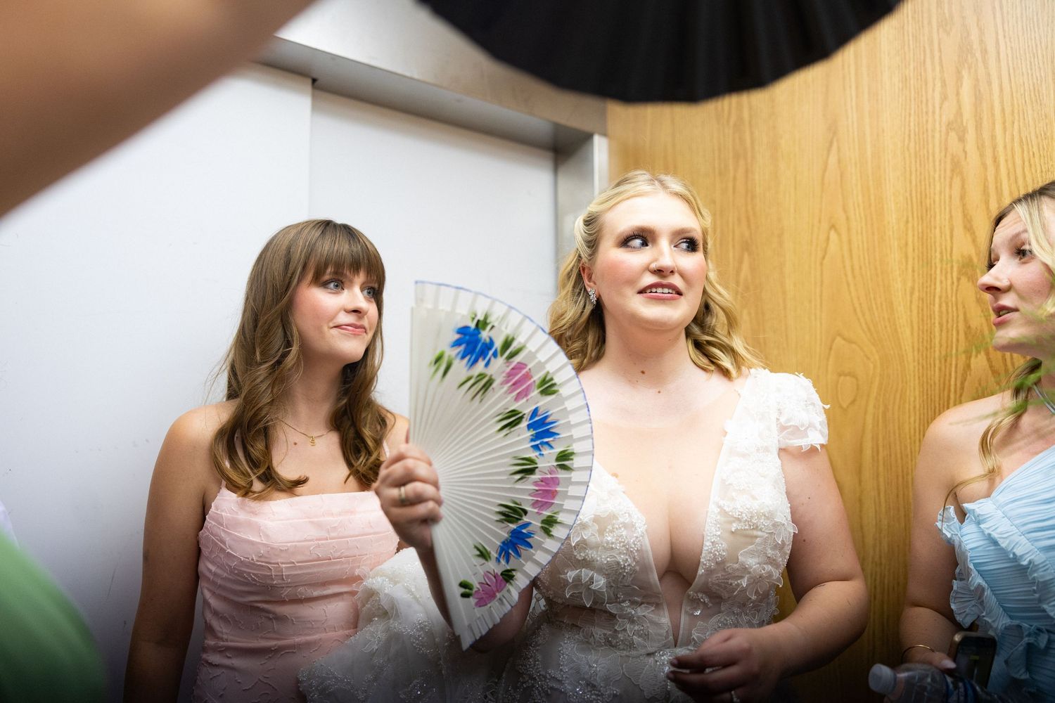 Bride in white lace gown holding floral fan with bridesmaids in pink dresses before wedding ceremony.