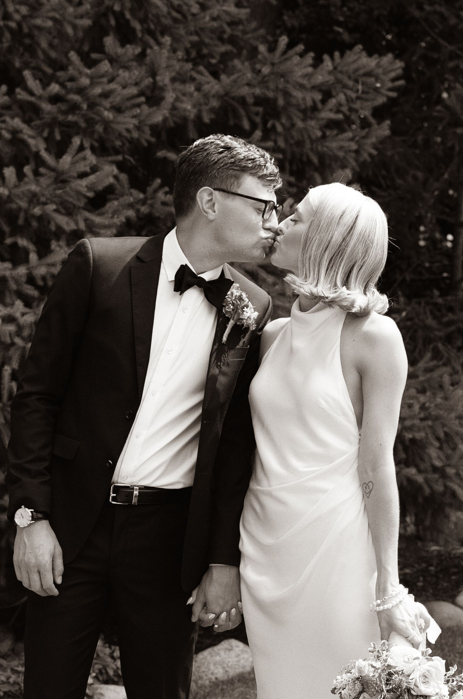 Black and white photo of newlyweds kissing, groom in tuxedo and bride in sleek white dress.
