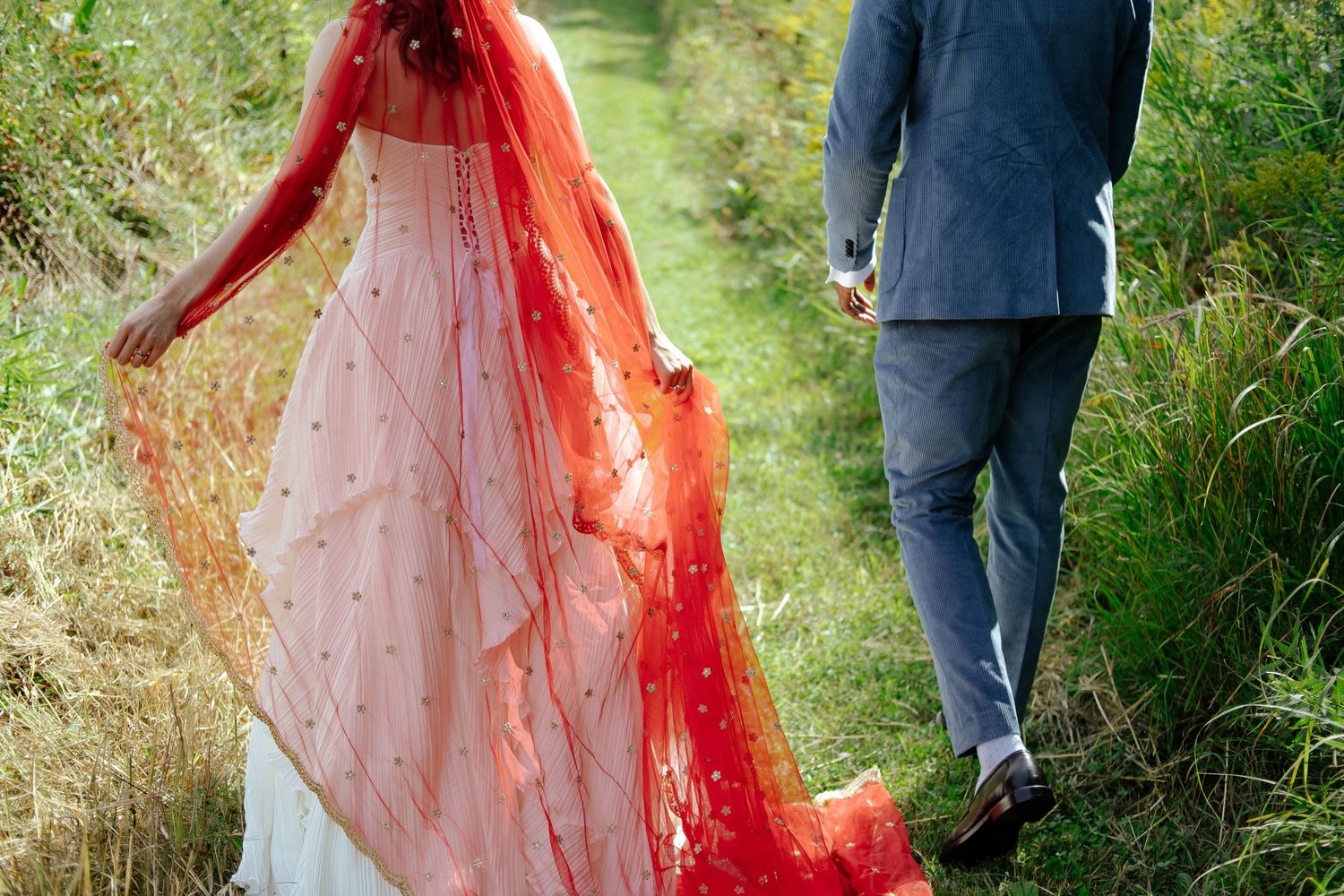Bride in white gown with red dupatta walks with groom in blue suit along a lush green garden path.