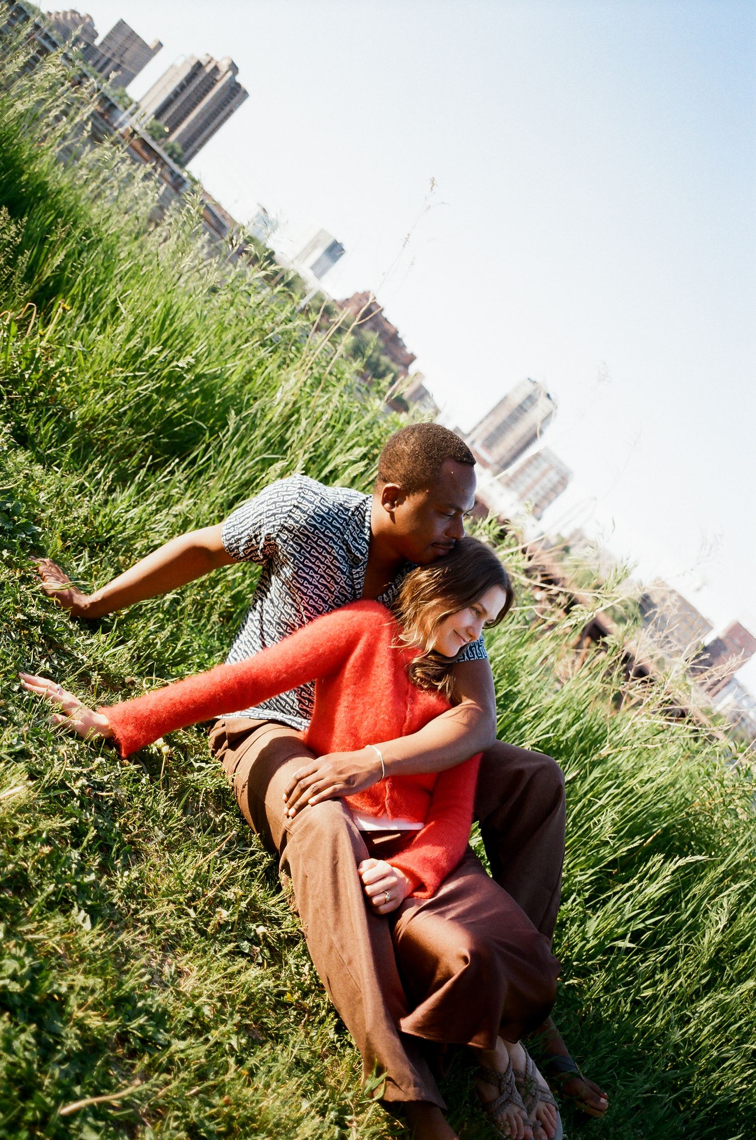 Couple relaxing together in tall green grass with urban cityscape in background.
