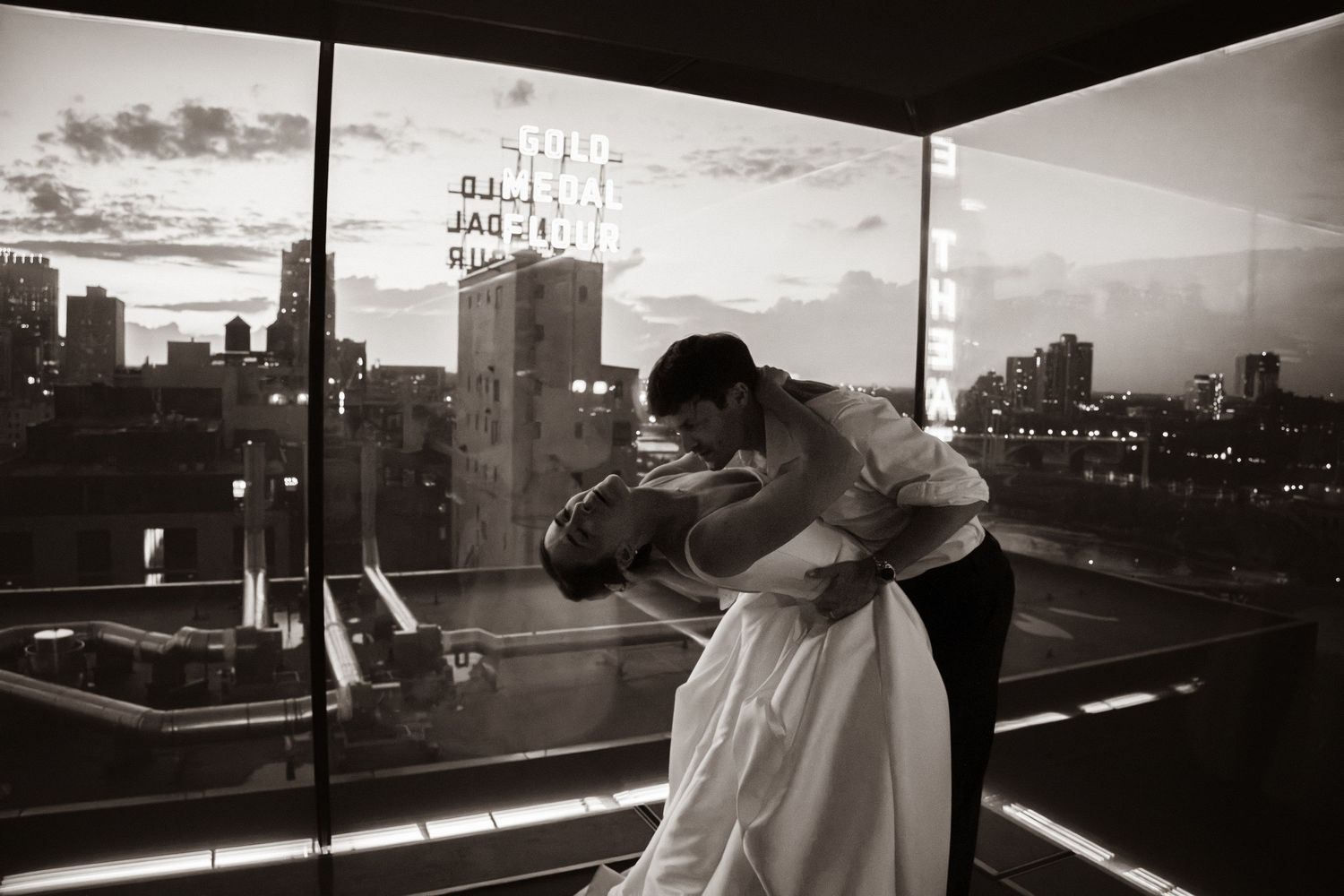 Black and white photo of a couple sharing a romantic dip kiss on a rooftop at dusk with city skyline backdrop.