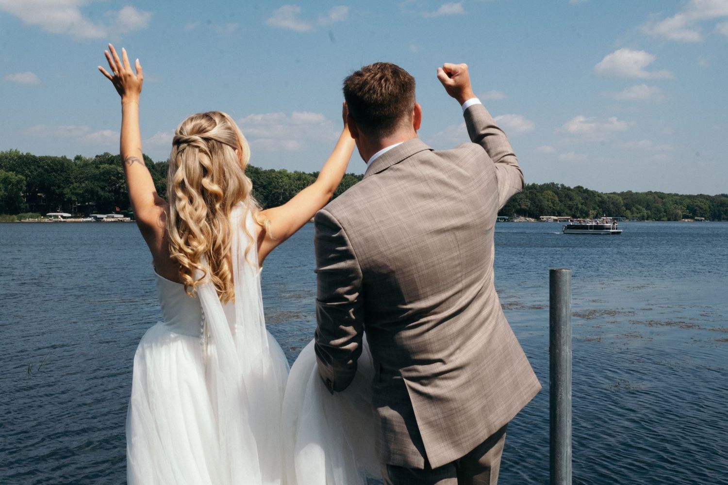 Bride and groom celebrating on a lakeside dock, raising their arms in joy on a sunny wedding day.
