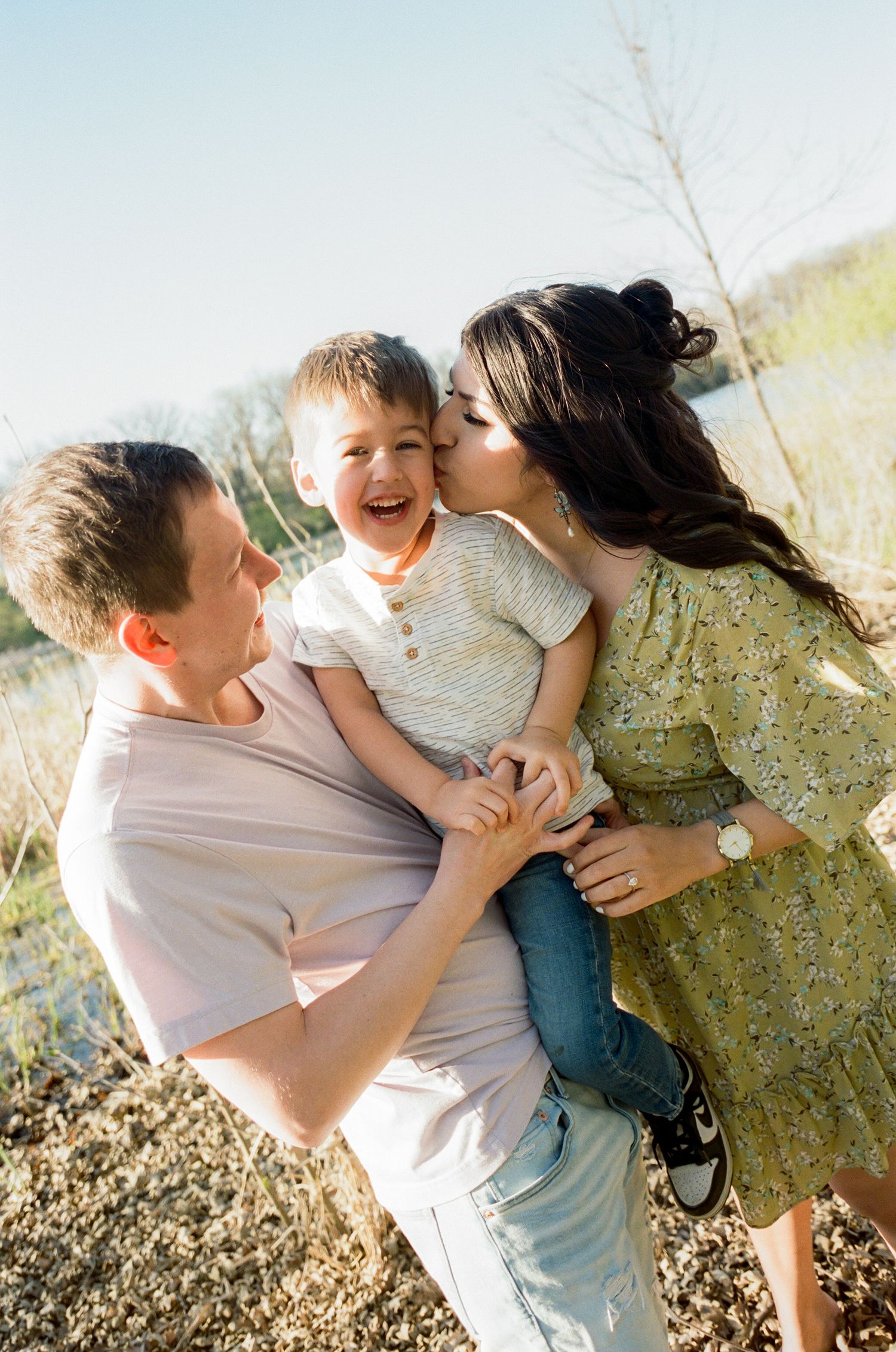 Happy family with laughing toddler boy being kissed by mother while father holds child.