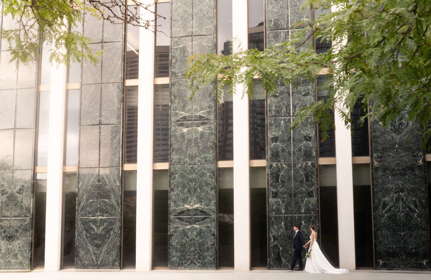 Bride and groom share a romantic moment beside a stunning green marble building facade with tall columns.