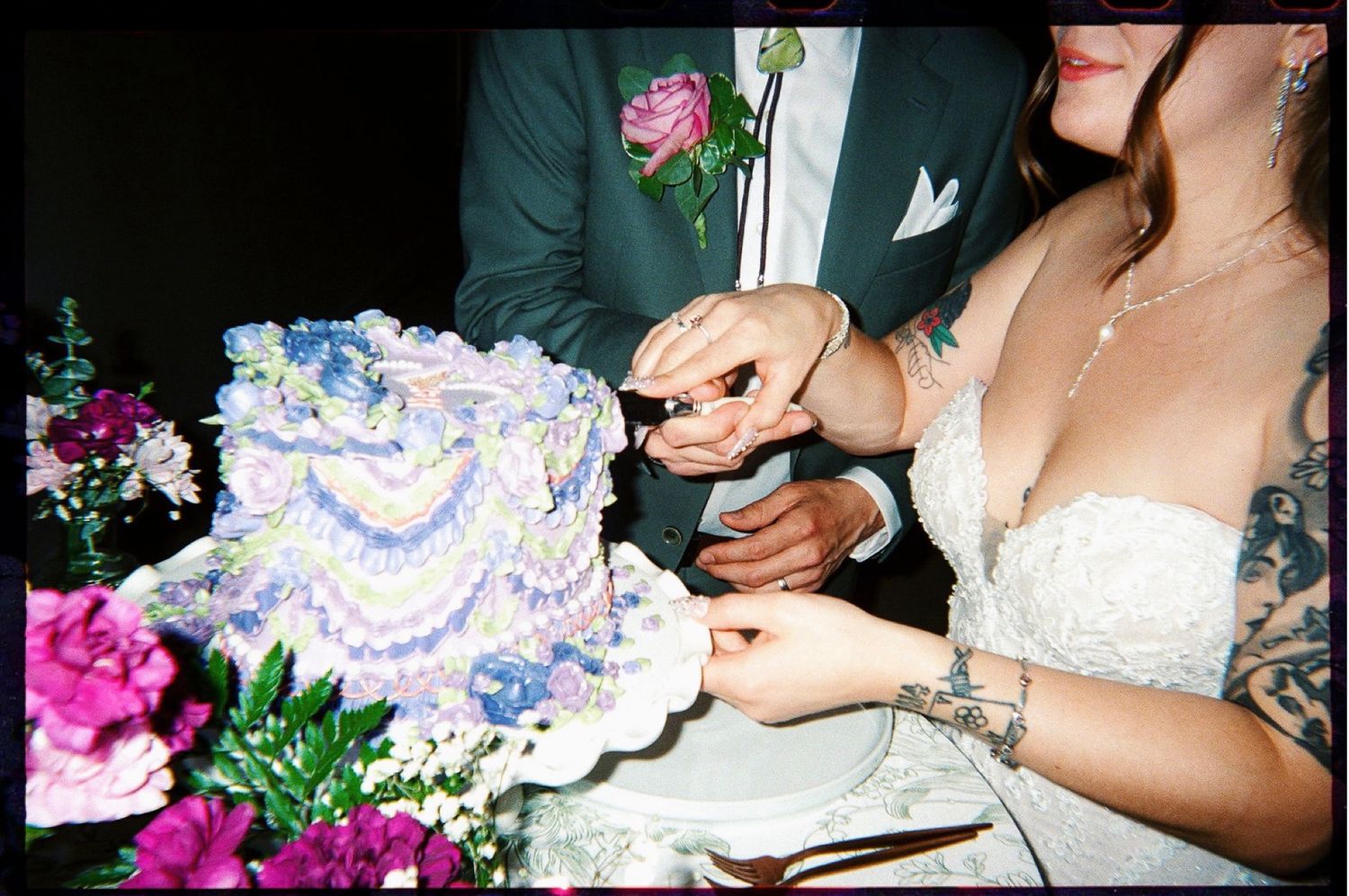 Bride and groom cutting a decorative blue and purple wedding cake surrounded by pink flowers.