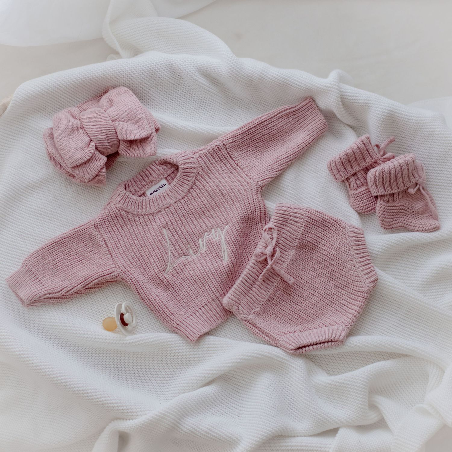 Pink knitted baby outfit with matching bow accessory and booties laid out on white fabric background.