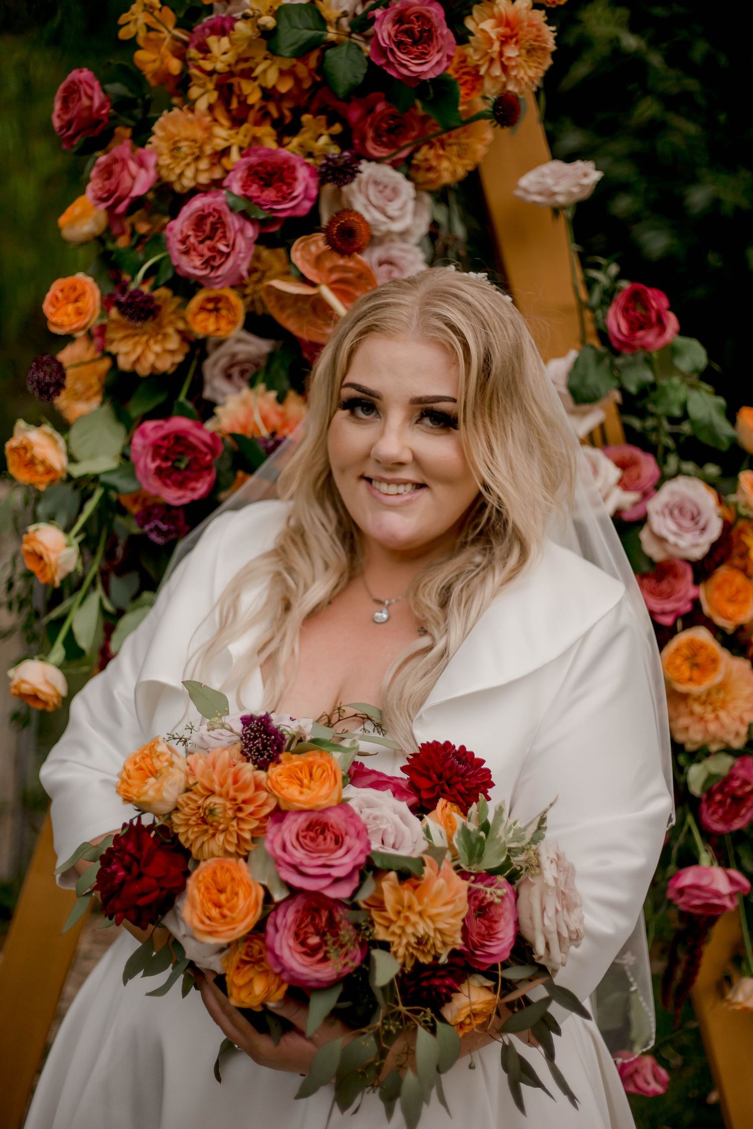 A bride in a white wedding dress holds a vibrant autumn-colored bouquet against a backdrop of roses and orange florals.