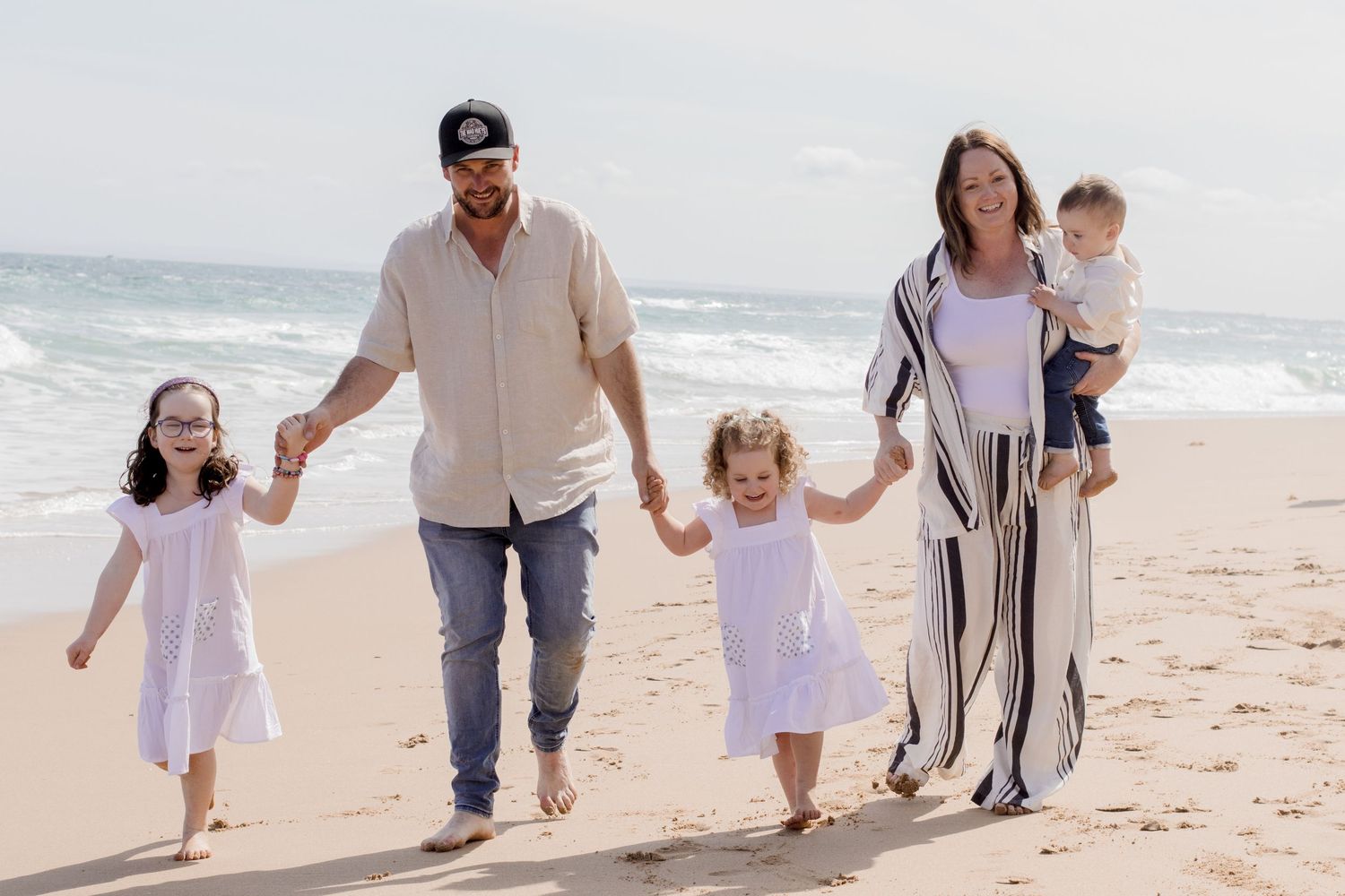 Family walking hand in hand along the shoreline wearing coordinated light colored outfits.