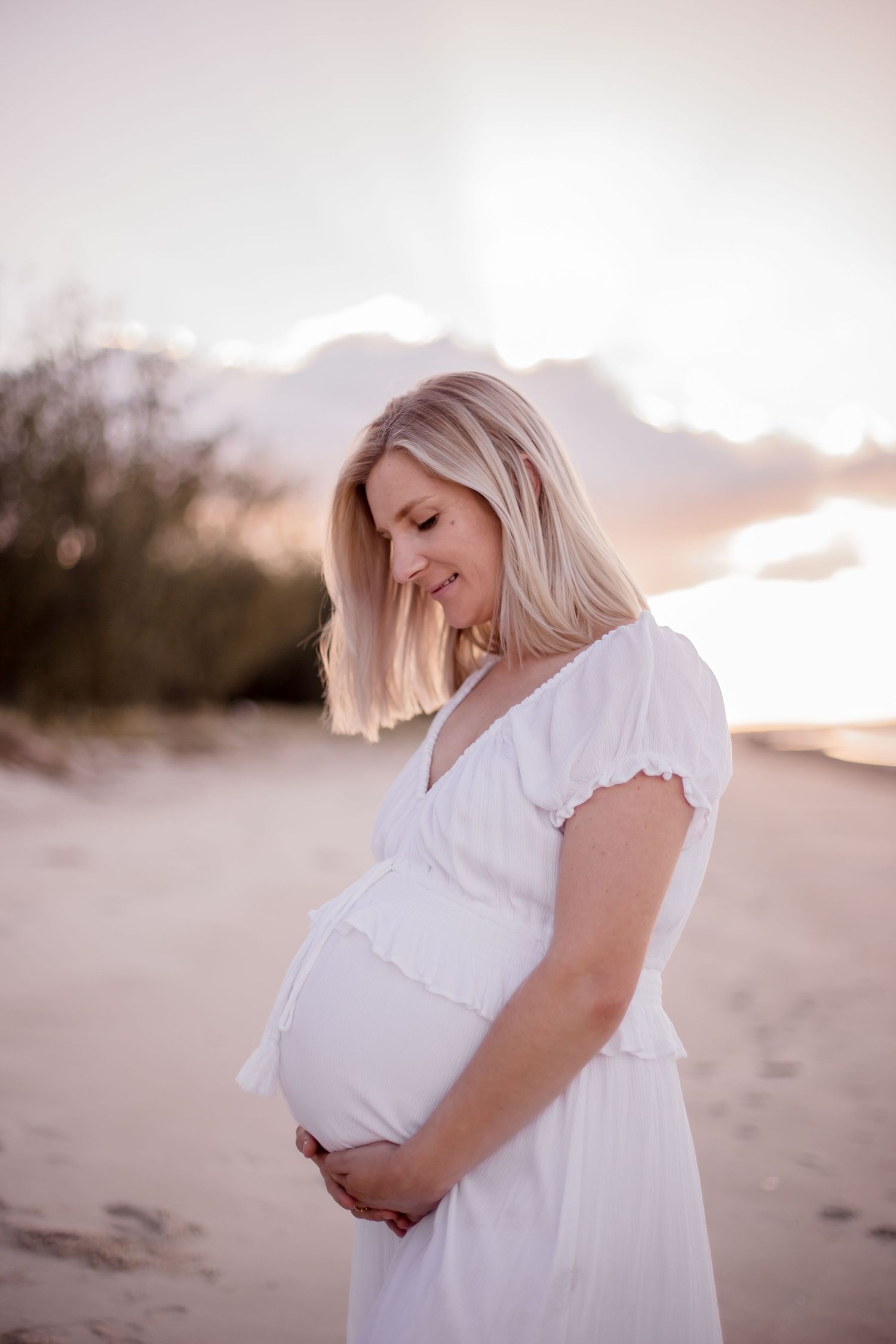 Expectant mother in white dress smiling while cradling her baby bump on beach at sunset.