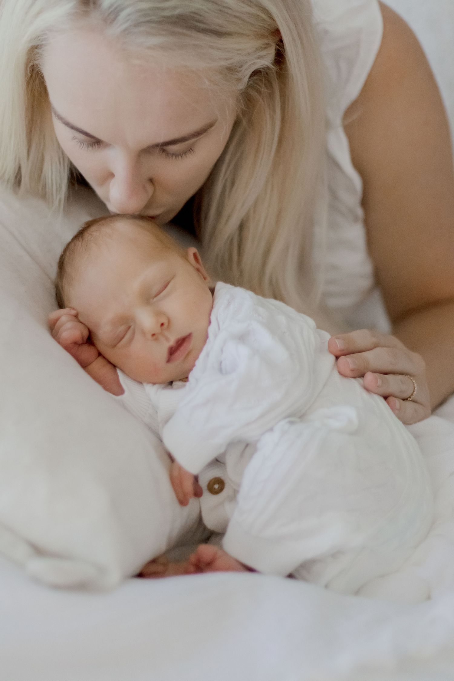 Mother gently kissing her sleeping newborn baby dressed in white.
