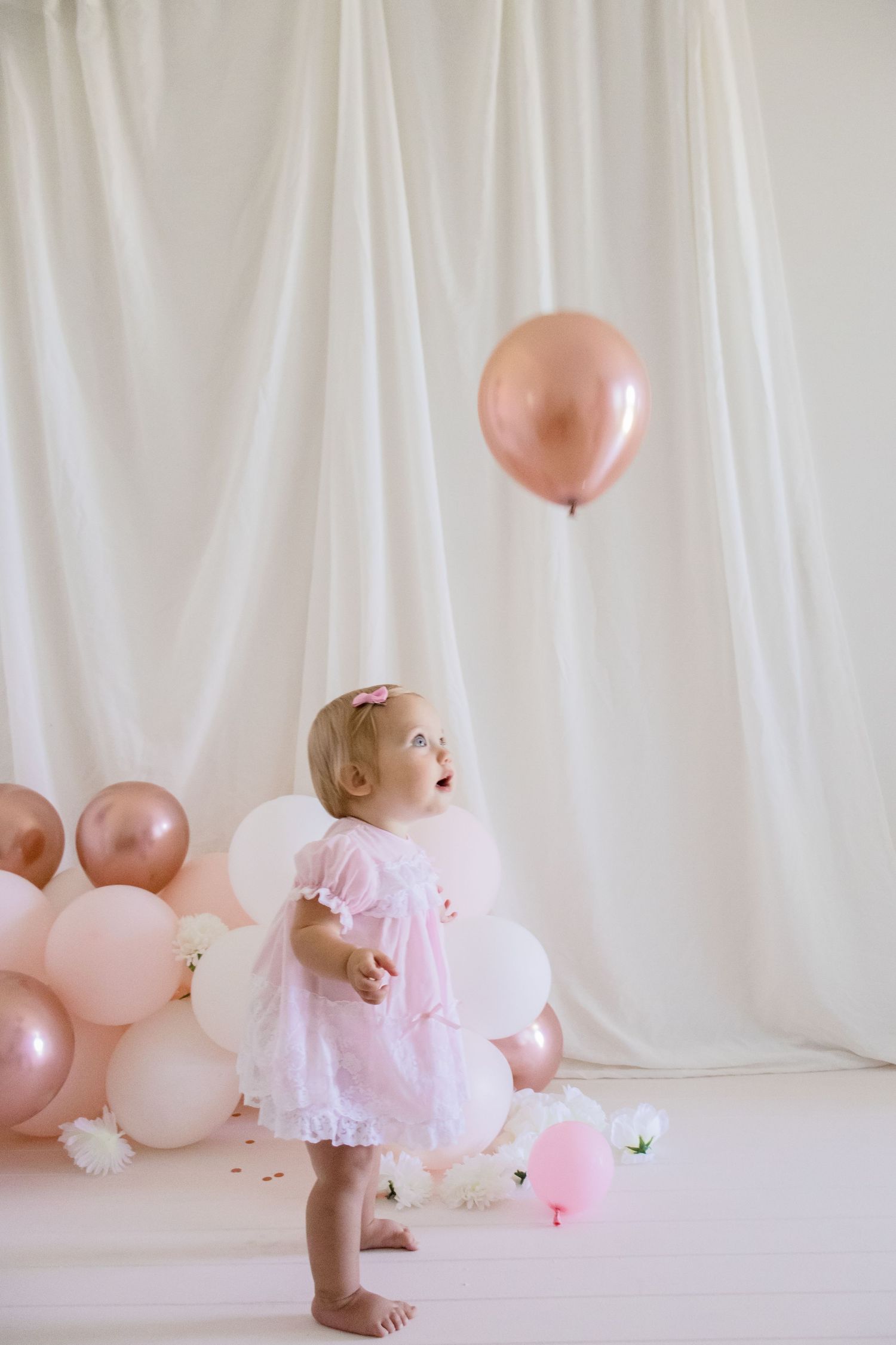 Baby in pink lace dress standing among floating pink balloons against white background.