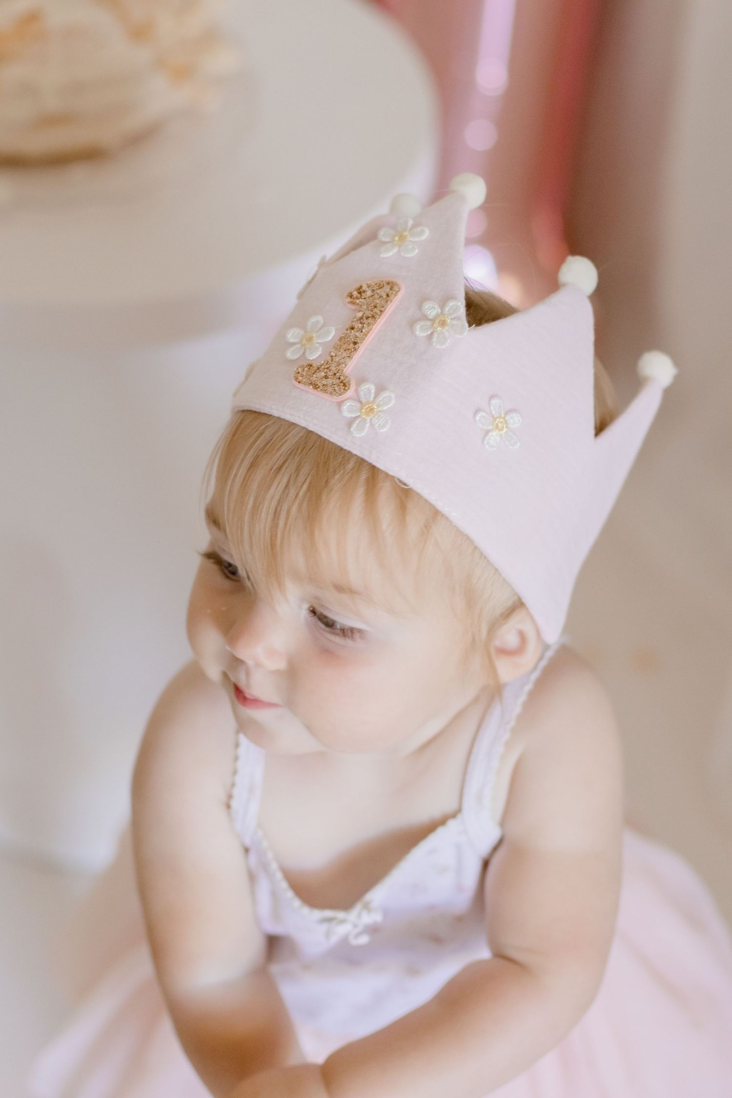 Baby in crown and white dress looking down during first birthday photoshoot.